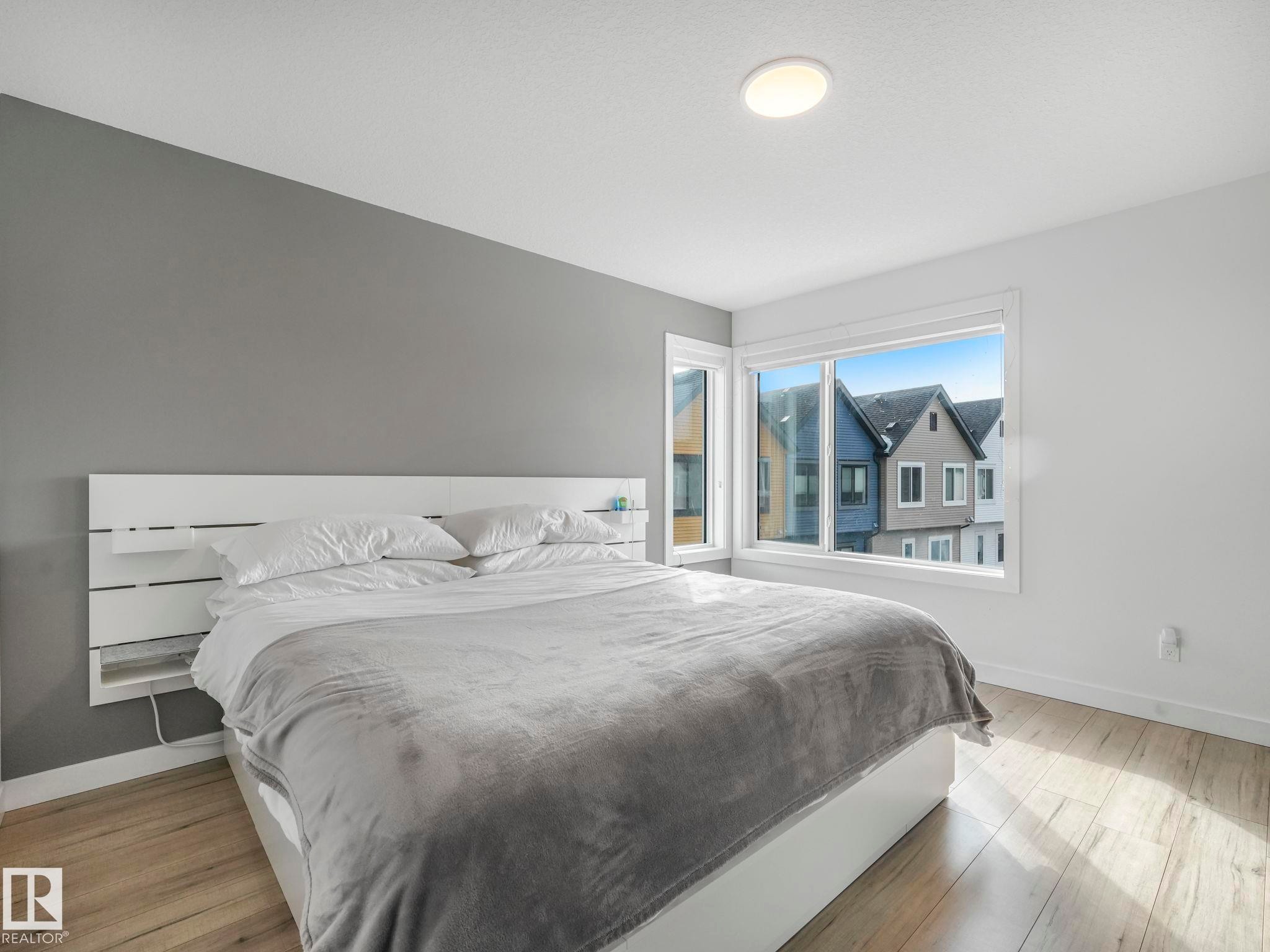 This bright bedroom features a modern white bed frame with integrated shelving, light wood flooring, and a large window offering views of the surrounding neighborhood properties - 95 1010 Rabbit Hill Road, Edmonton, AB - Indoor Photo Showing Bedroom
