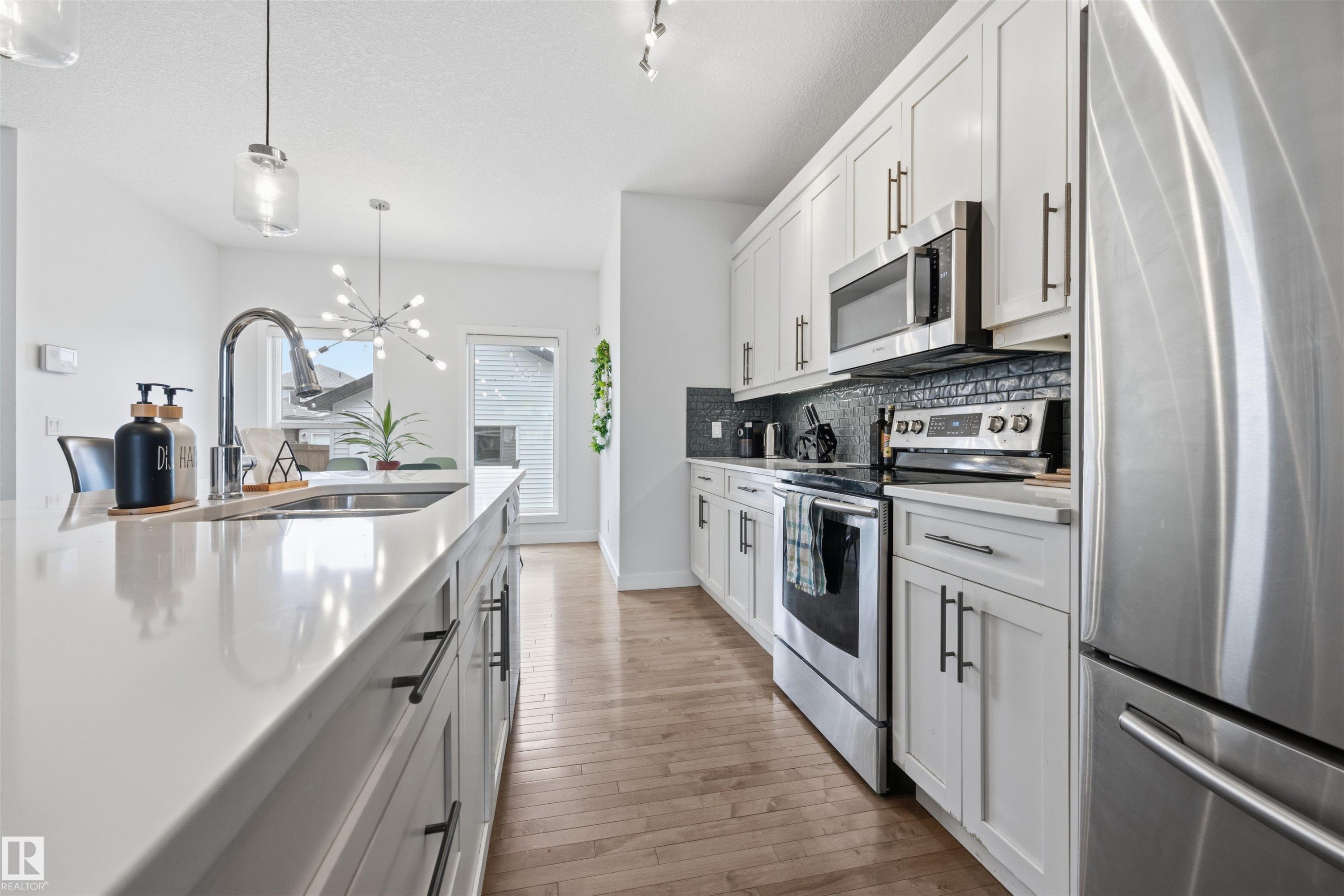 The kitchen features an island with an integrated sink, white cabinetry with dark hardware, stainless steel appliances, and wood flooring - 7123 Cardinal Way, Edmonton, AB - Indoor Photo Showing Kitchen With Double Sink With Upgraded Kitchen