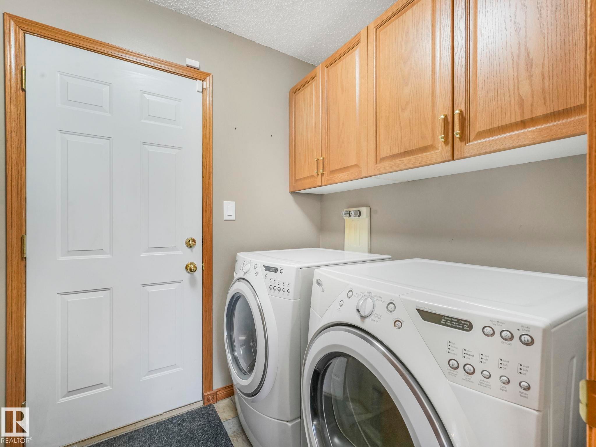 Dedicated laundry area featuring overhead wooden cabinetry, a white paneled door with brass hardware, and two front-loading appliances - 11704 11 Avenue, Edmonton, AB - Indoor Photo Showing Laundry Room