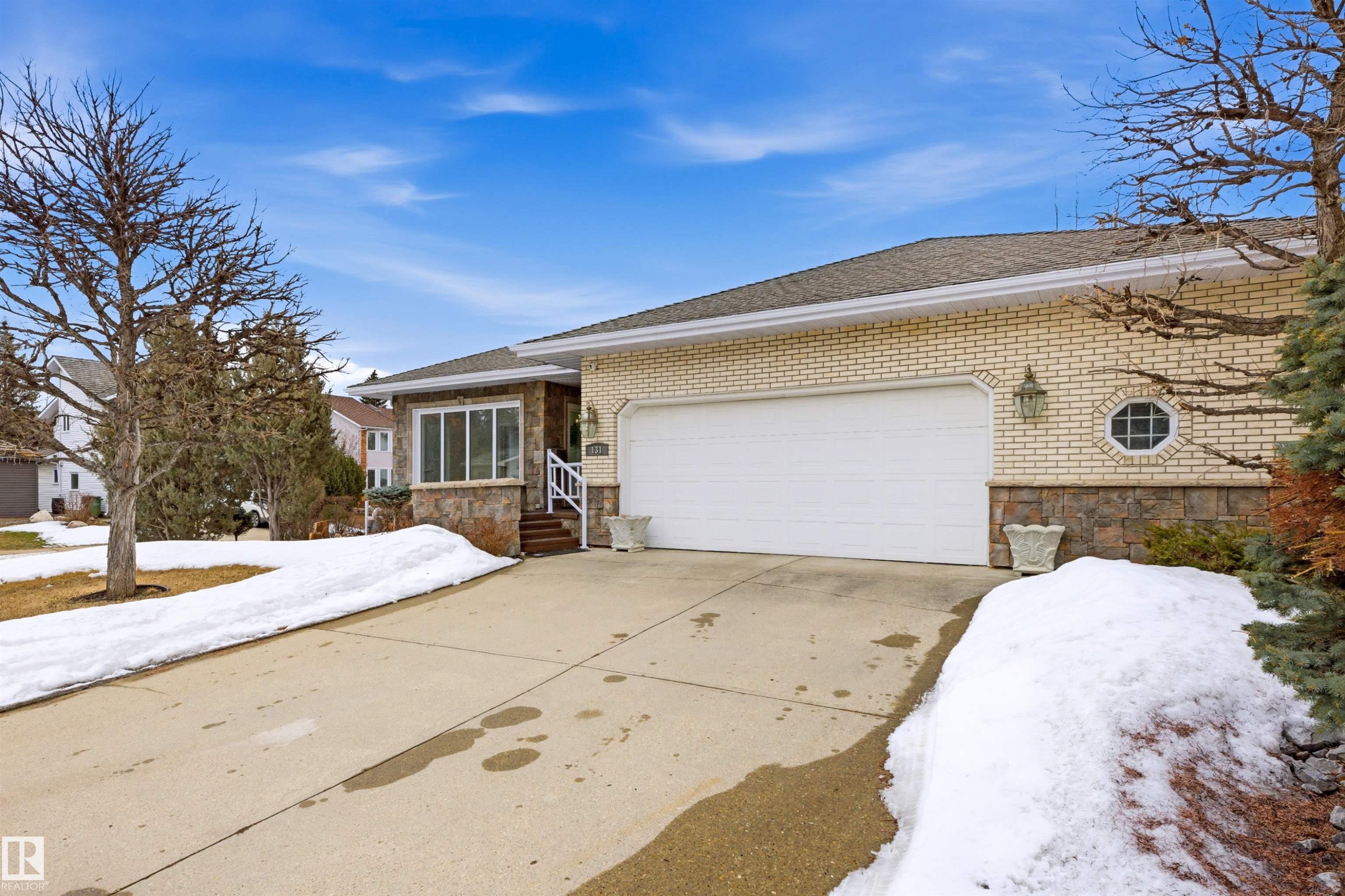 This property features a light-colored brick exterior with stone accents, a two-car garage with a white door, and a concrete driveway - 131 Walsh Crescent, Edmonton, AB - Outdoor