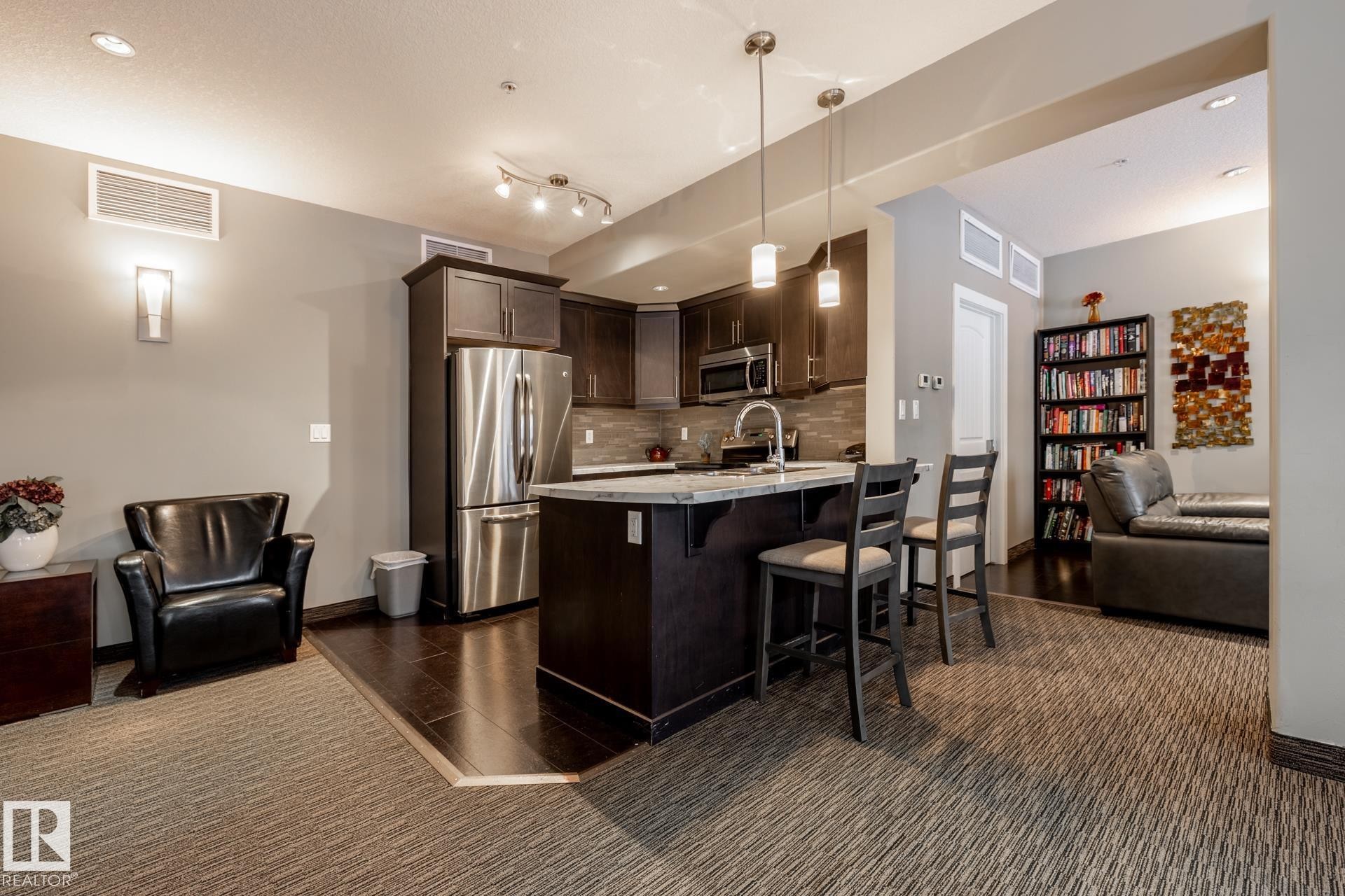 The property features a kitchen with dark cabinetry, stainless steel appliances, and a central island with bar seating - 454 6079 Maynard Way, Edmonton, AB - Indoor Photo Showing Kitchen With Upgraded Kitchen