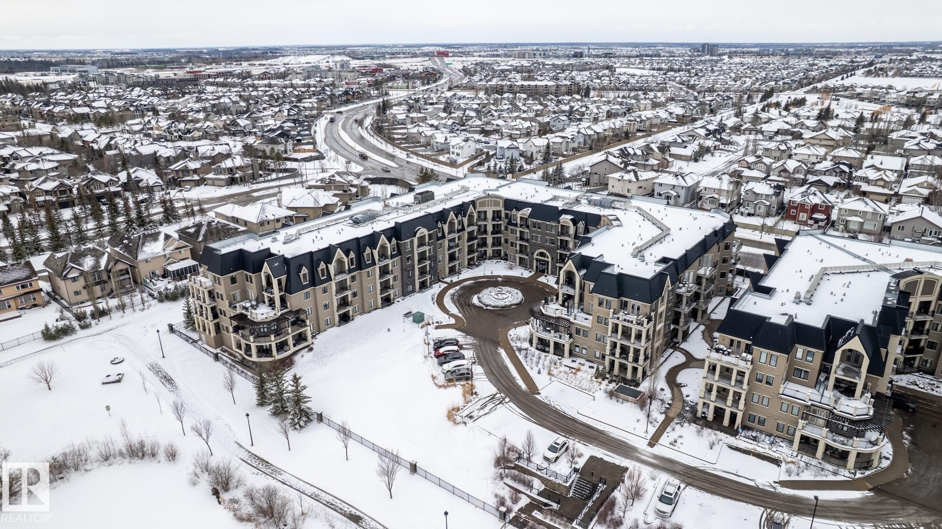 A modern building with a distinctive dark roof, showcasing several balconies and a central driveway entrance - 454 6079 Maynard Way, Edmonton, AB - Outdoor With View