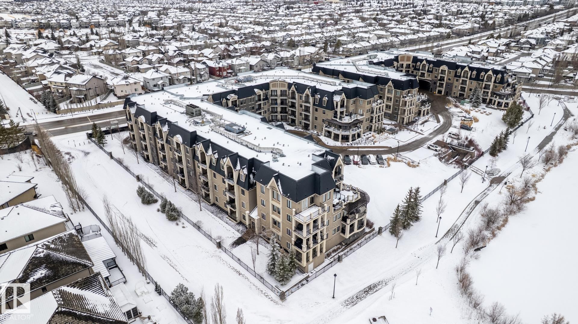 This property features a multi-story building with a distinctive dark roof and light-colored facade, surrounded by a snow-covered landscape - 454 6079 Maynard Way, Edmonton, AB - Outdoor