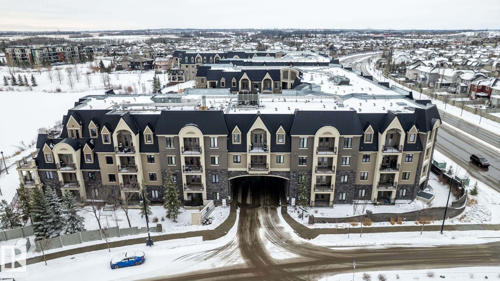 The property features a multi-story building with a distinctive facade combining stone and stucco, topped with dark sloped roofs and dormer windows - 454 6079 Maynard Way, Edmonton, AB - Outdoor With Facade