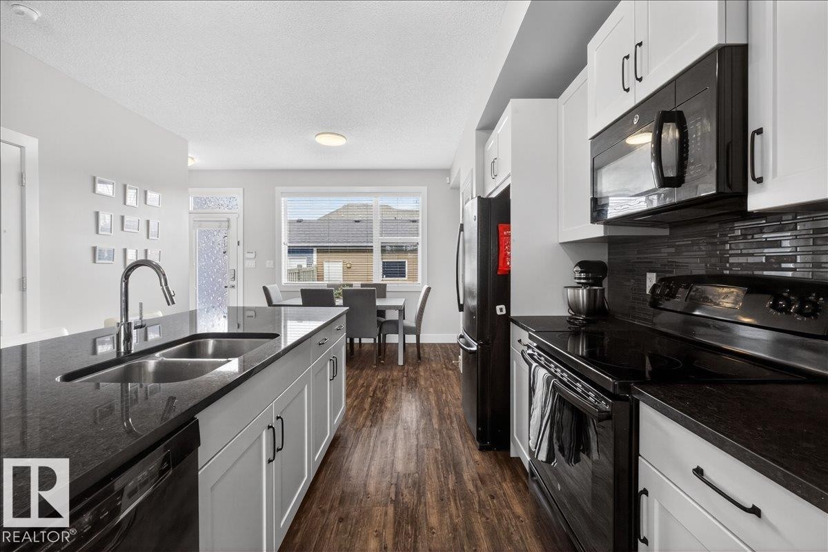 The kitchen features an island with a double basin sink and a modern faucet, dark countertops, white cabinetry, and black appliances - 9034 Rosenthal Link, Edmonton, AB - Indoor Photo Showing Kitchen With Double Sink With Upgraded Kitchen