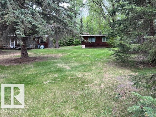 Expansive grassy yard area with mature evergreen trees, featuring a dark-sided structure with multiple windows and a separate smaller dwelling with a covered porch - 1040 &1040 A&B Mitchell Beach Beach, Rural Leduc County, AB