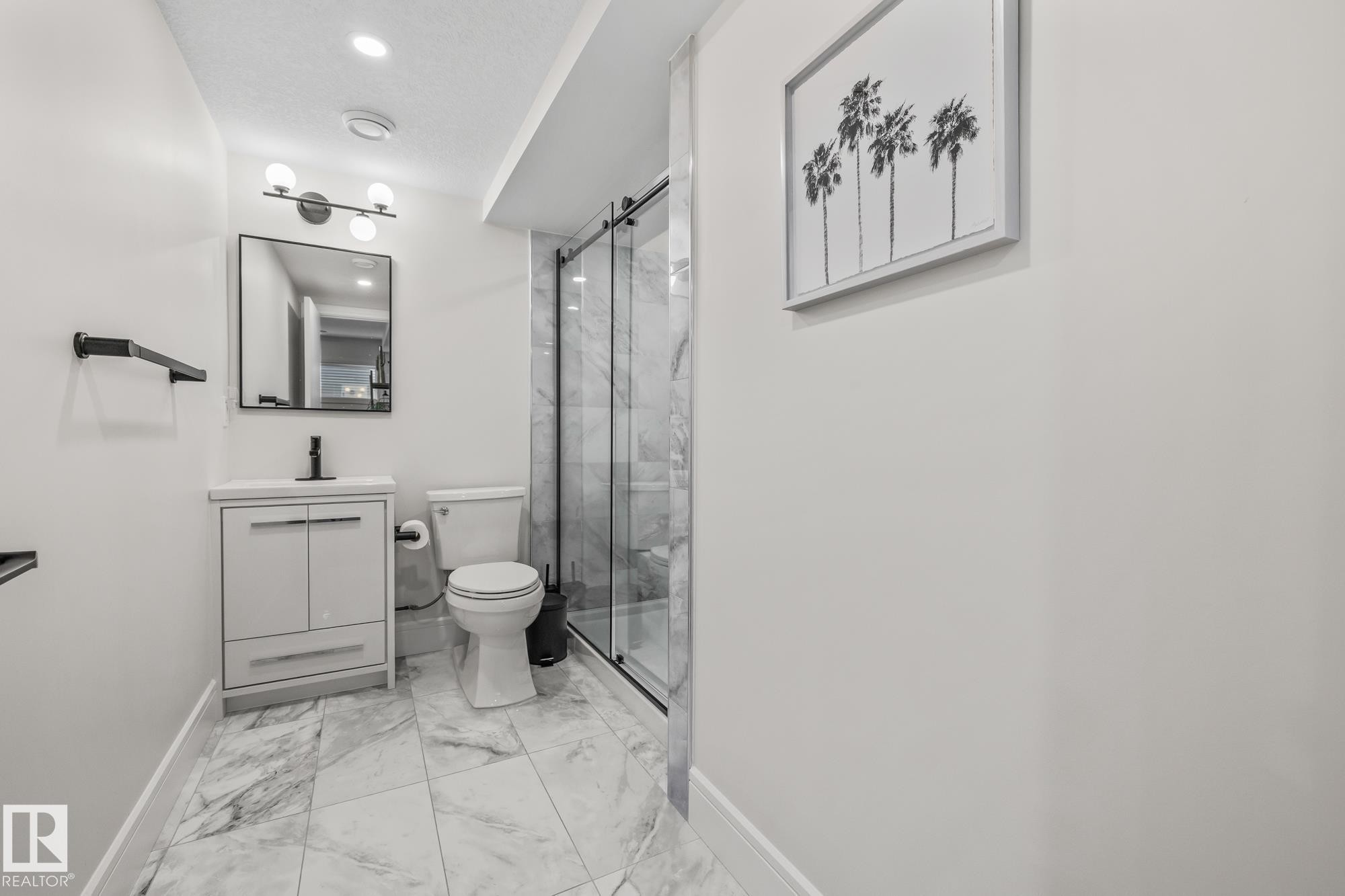 This bathroom features a contemporary vanity with a white countertop and dark-toned faucet, a glass-enclosed shower with marble-patterned walls, and a toilet - 3864 Robins Crescent, Edmonton, AB - Indoor Photo Showing Bathroom
