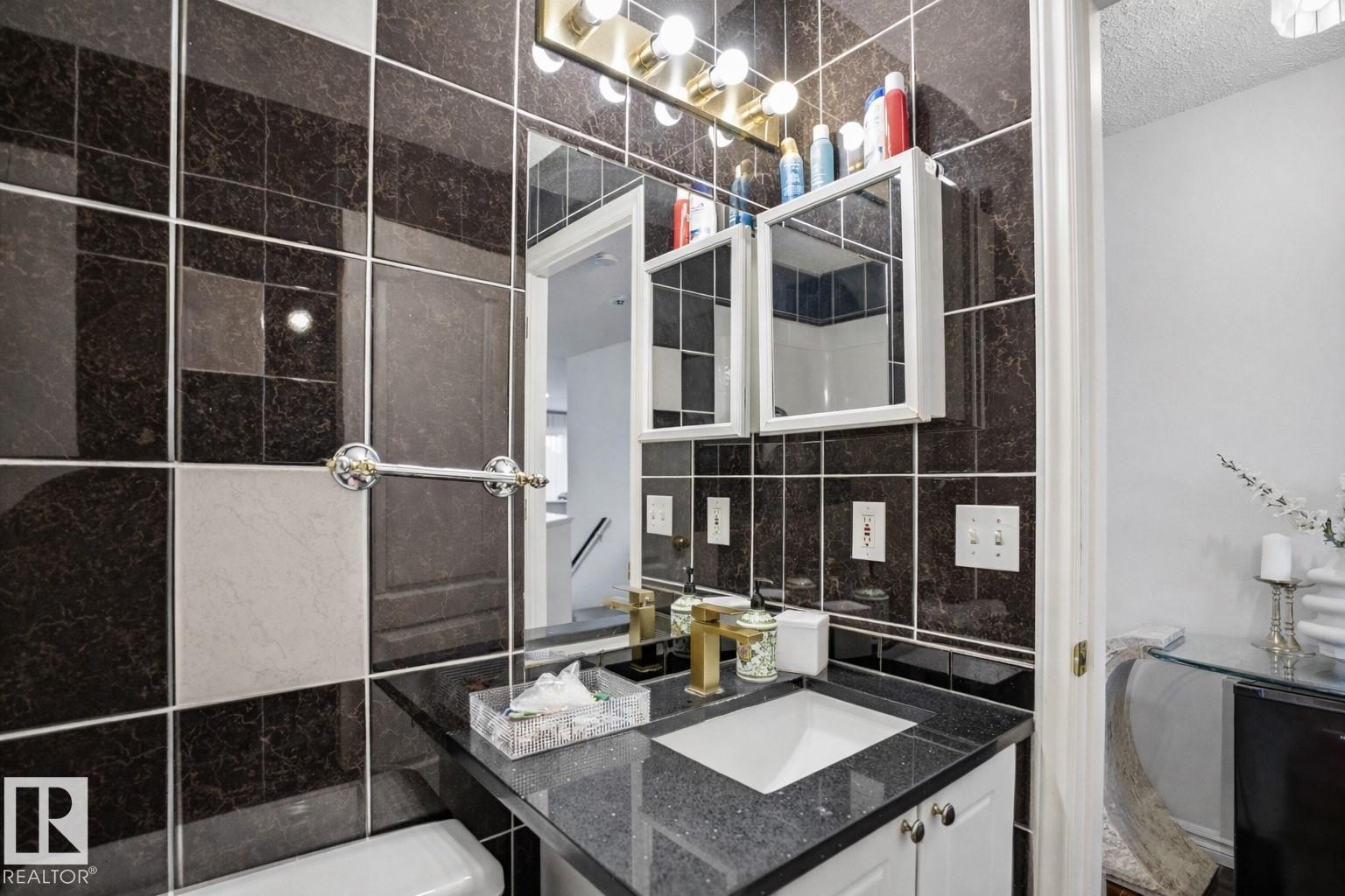 Bathroom featuring dark tiled walls with light accent tiles, a vanity with a dark speckled countertop, and a gold-toned faucet - 4619 202 Street, Edmonton, AB - Indoor Photo Showing Bathroom