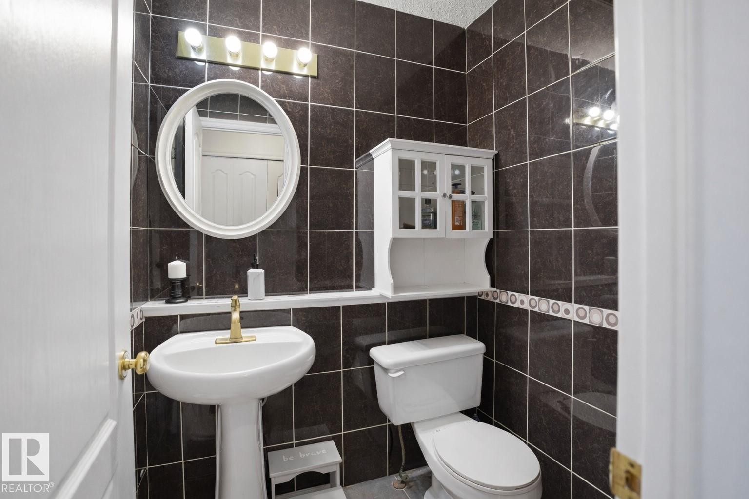 The bathroom features dark tiled walls, a white pedestal sink with a brass-toned faucet, and a wall-mounted mirror with a white frame - 4619 202 Street, Edmonton, AB - Indoor Photo Showing Bathroom