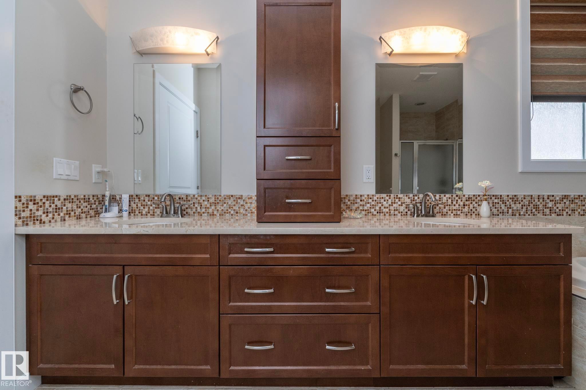 This bathroom features a double vanity with brown cabinetry, a light-colored countertop, and a mosaic tile backsplash - 307 Ravine Close, Devon, AB - Indoor Photo Showing Bathroom