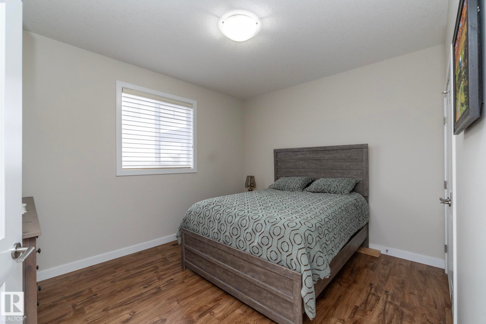 The room features hardwood style flooring and a window with blinds, allowing for adjustable natural light - 307 Ravine Close, Devon, AB - Indoor Photo Showing Bedroom