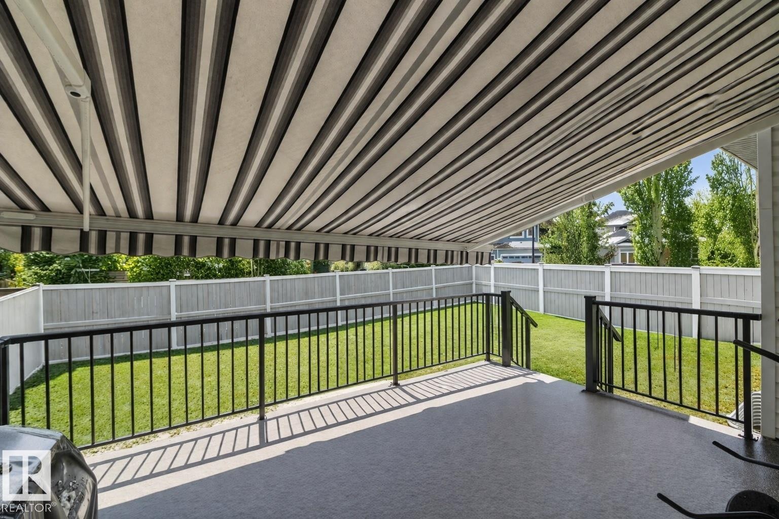 The property features an outdoor patio with a striped retractable awning, a black metal railing, and a fenced yard with a lawn - 307 Ravine Close, Devon, AB - Outdoor With Deck Patio Veranda
