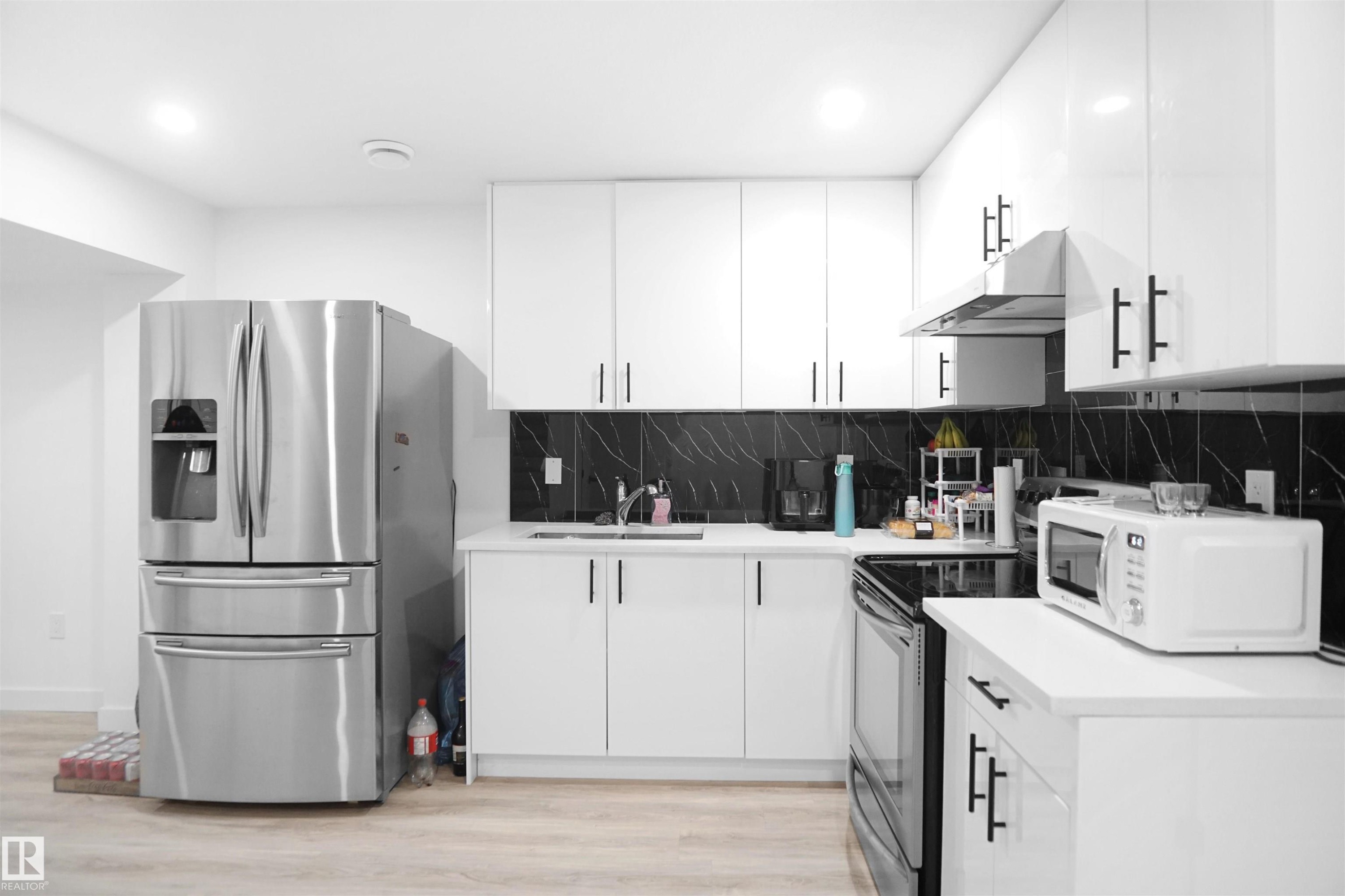 The kitchen features white cabinetry with black hardware, a stainless steel refrigerator, and a dark backsplash with white veining - Edmonton, AB - Indoor Photo Showing Kitchen