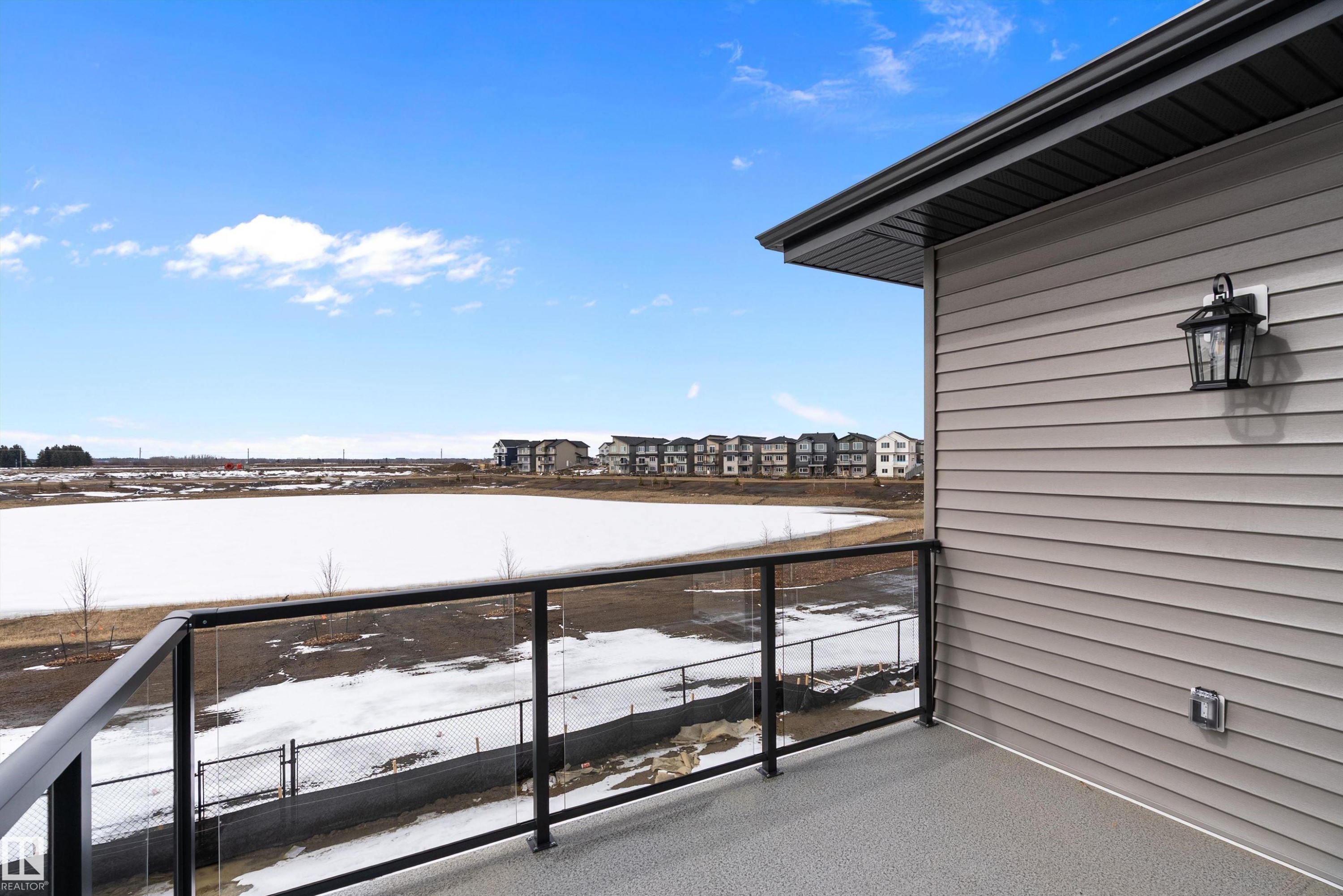 This property features a balcony with a black metal and glass railing, offering views of a frozen body of water and distant buildings - 219 Crystal Creek Dr, Leduc, AB - Outdoor With Exterior