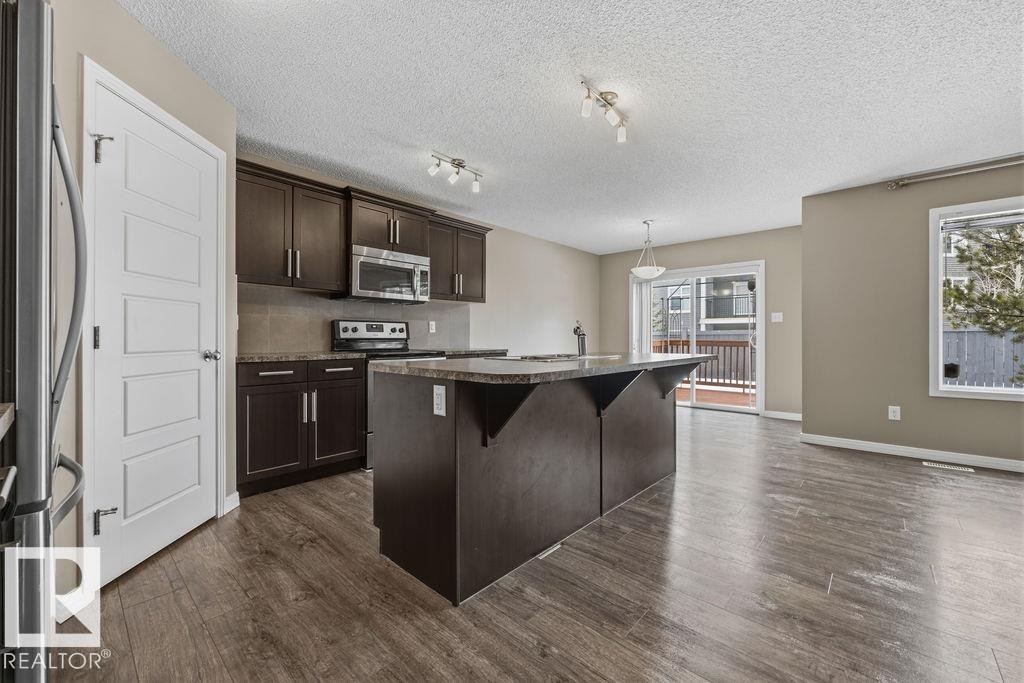 Spacious open-concept kitchen featuring dark wood cabinetry, stainless steel appliances, a large island with a sink, and light-colored flooring - 7873 170A Avenue, Edmonton, AB - Indoor Photo Showing Kitchen With Stainless Steel Kitchen With Upgraded Kitchen