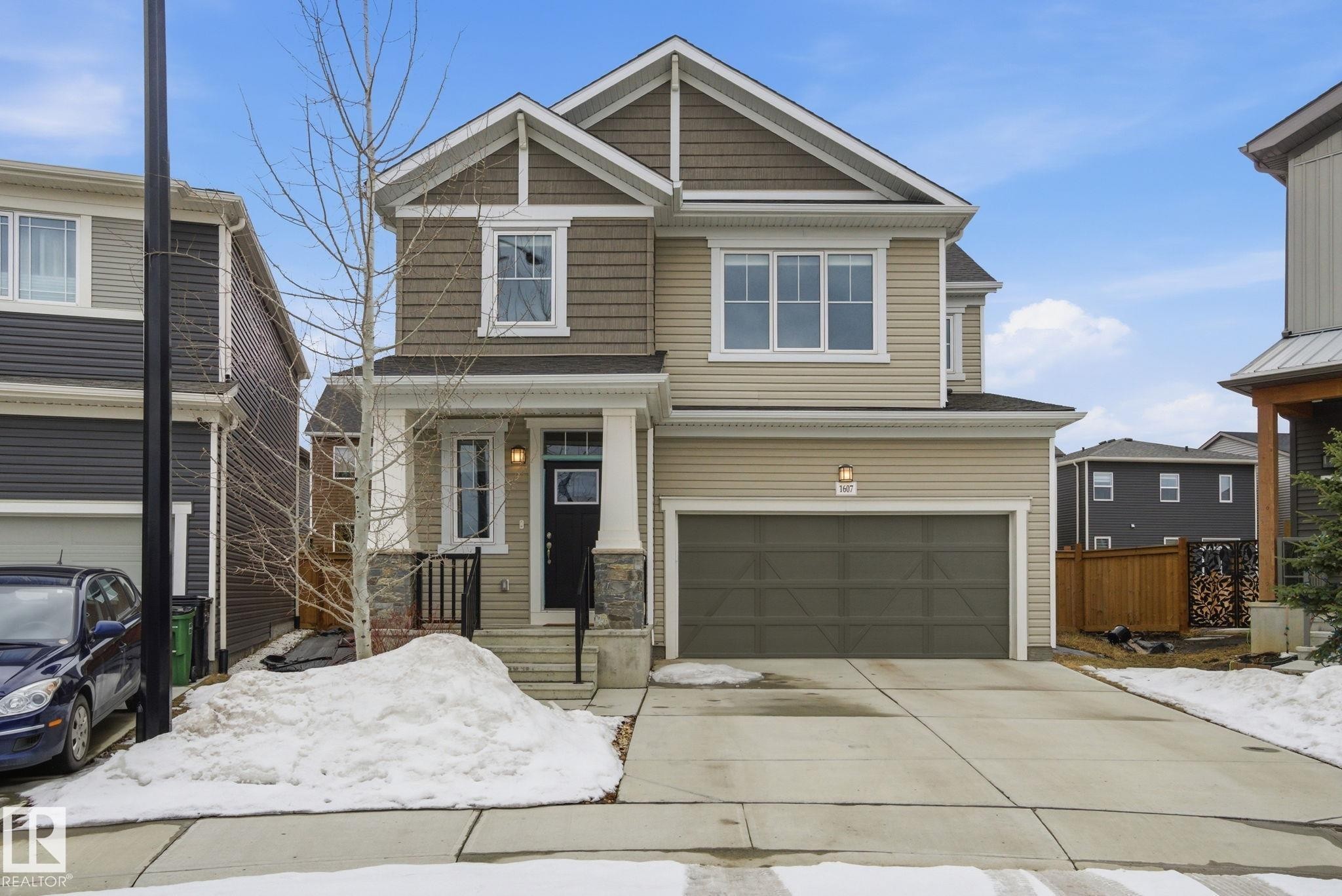 The property features a two-story exterior with light beige and brown siding, a covered front entrance with a black door, and an attached two-car garage - 1607 202 Street, Edmonton, AB - Outdoor With Facade