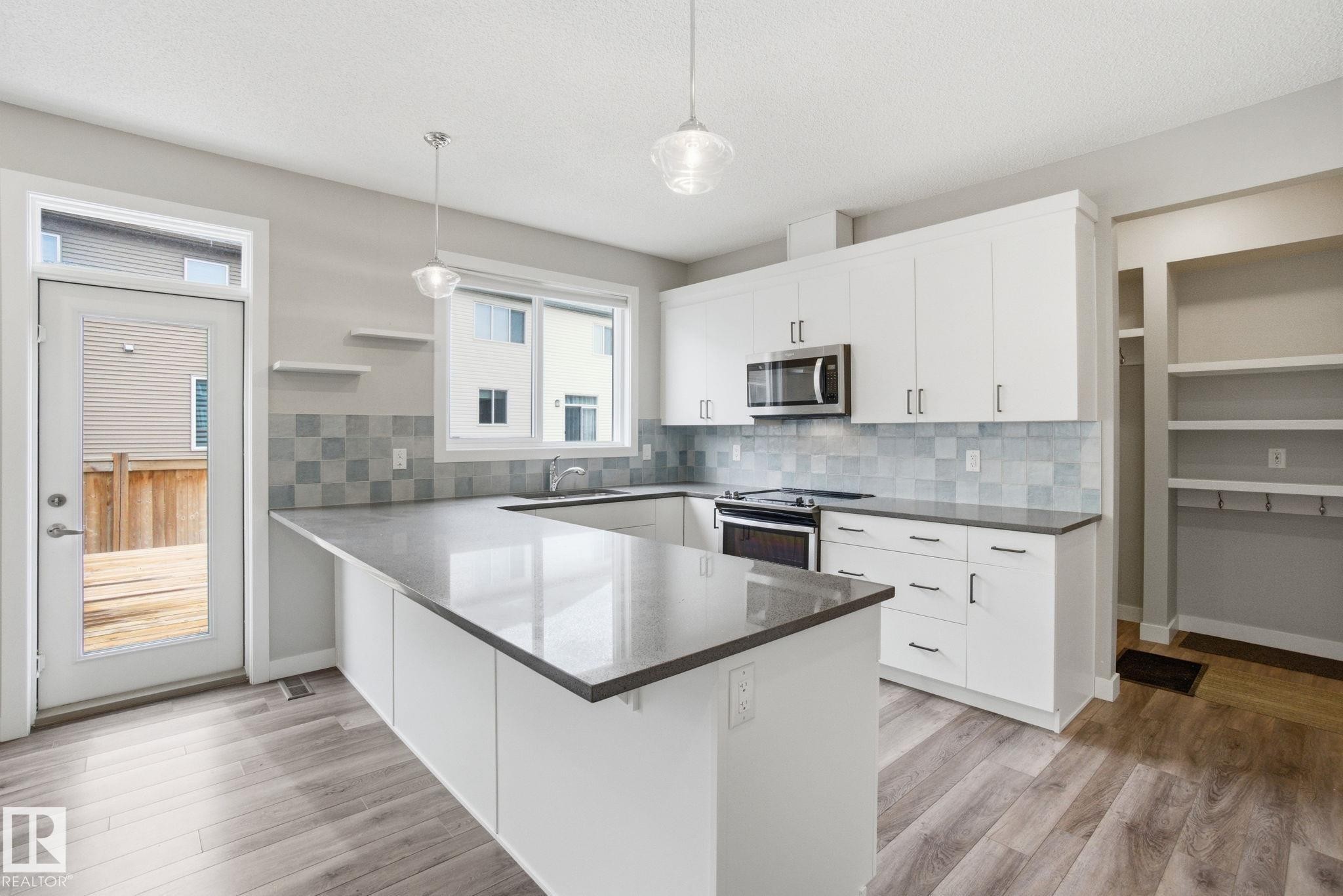Modern kitchen featuring white cabinetry, a stainless steel microwave and oven, a grey tiled backsplash, a kitchen island with a grey countertop, and light wood-style flooring - 1607 202 Street, Edmonton, AB - Indoor Photo Showing Kitchen With Upgraded Kitchen