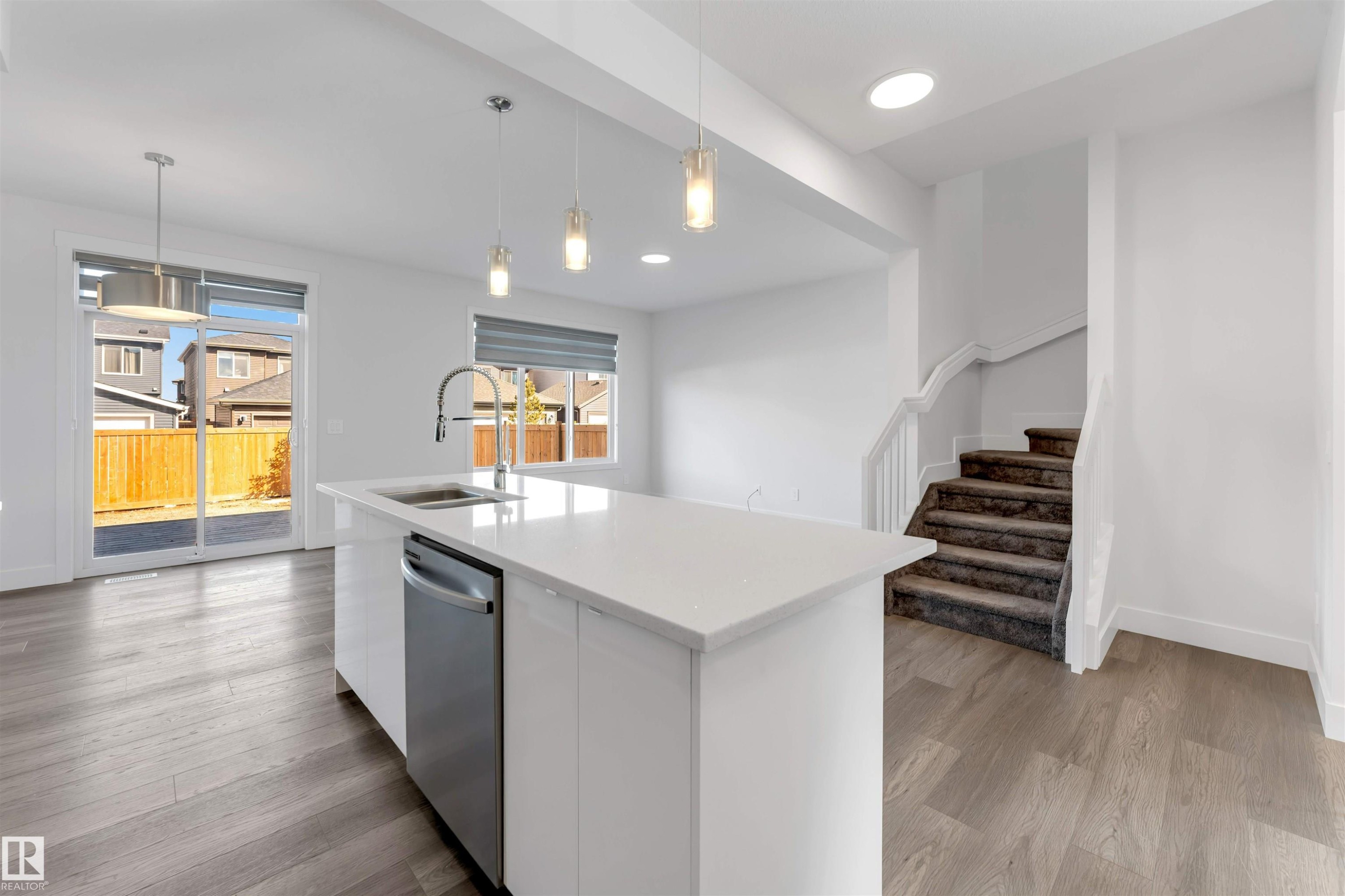 Open concept living area featuring light wood flooring, a kitchen island with a sink and stainless steel dishwasher, and a staircase with carpeted treads - 1663 Cavanagh Boulevard Sw, Edmonton, AB - Indoor Photo Showing Kitchen With Double Sink