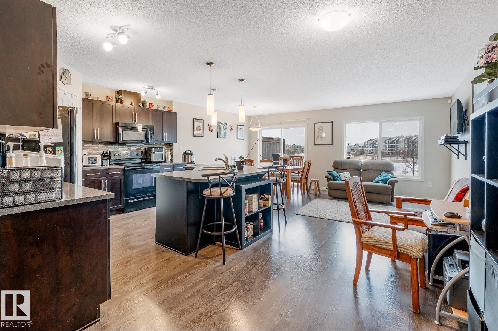 This open-concept living area features rich hardwood flooring, a well-appointed kitchen with dark cabinetry and stainless steel appliances, and an island with seating - 3109 Carpenter Landing, Edmonton, AB - Indoor Photo Showing Kitchen With Upgraded Kitchen