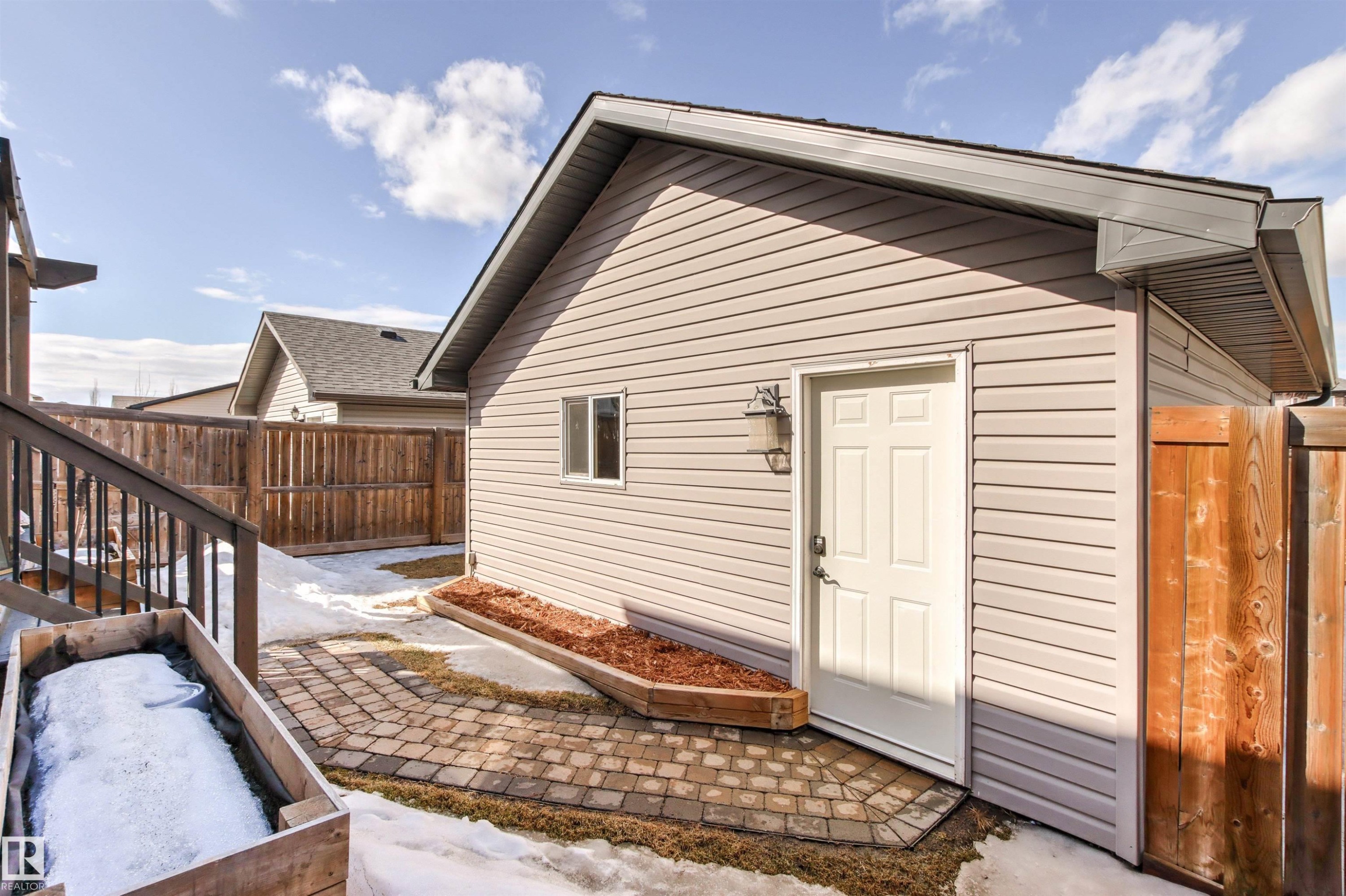 The property features a building with light-colored siding, a white door, and a small window, set on a paver patio - 38 Hewitt Circle, Spruce Grove, AB - Outdoor With Exterior