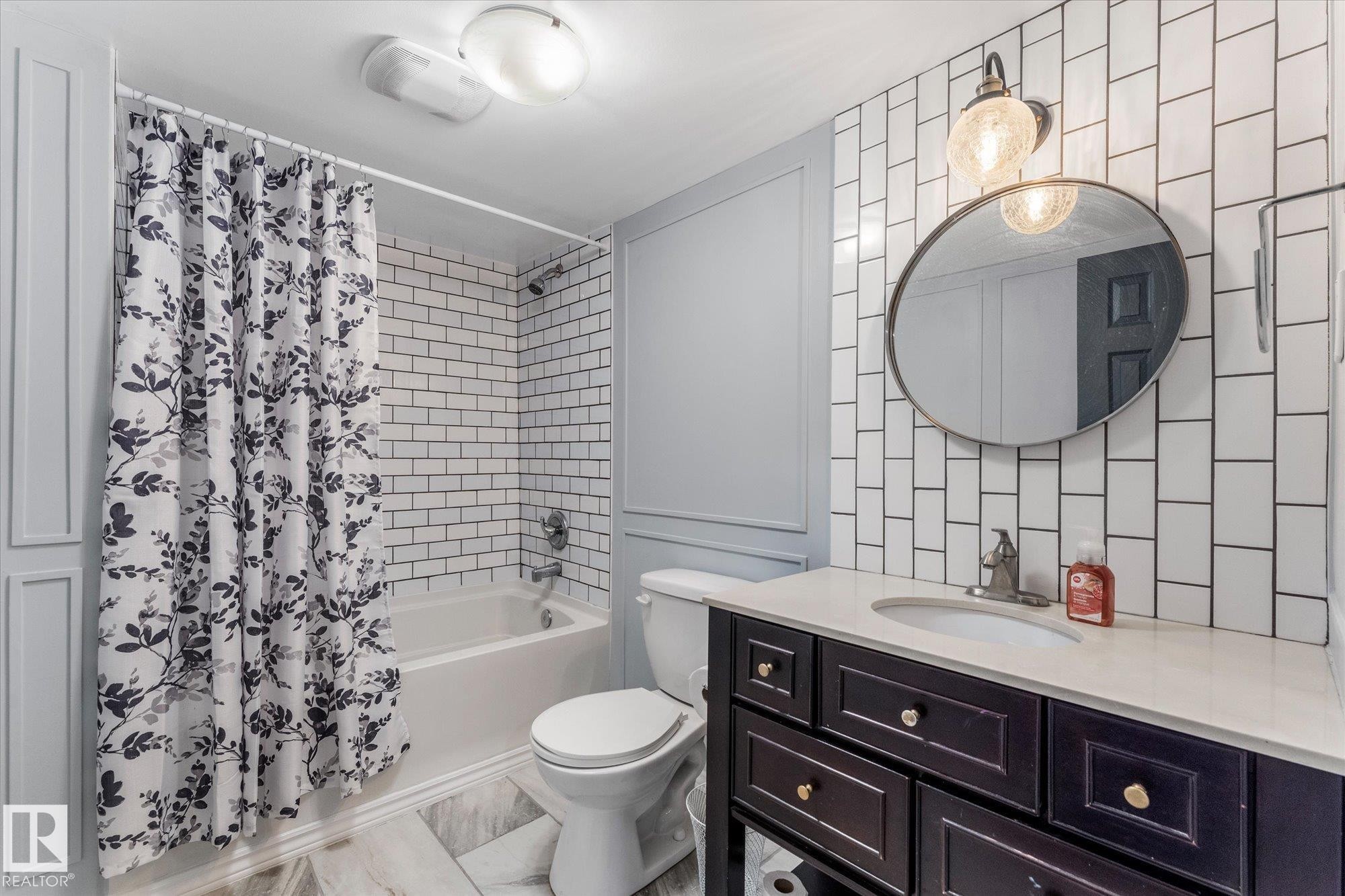 Bathroom featuring a subway tile shower surround, a full vanity with a white countertop and dark cabinetry, and an oval mirror with a wall-mounted light fixture - 2626 49 Street, Edmonton, AB - Indoor Photo Showing Bathroom