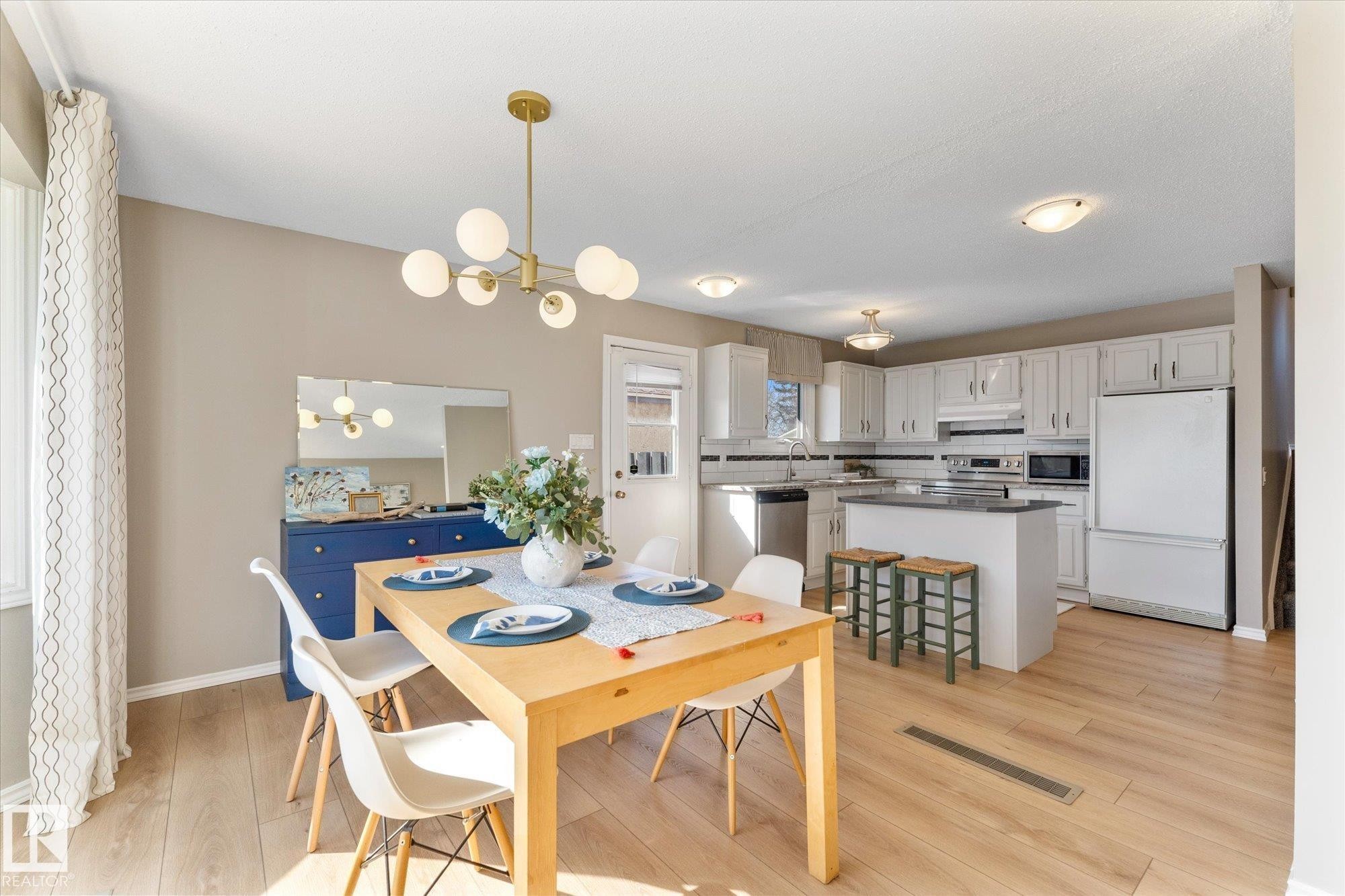 This open-concept area features hardwood flooring, a modern chandelier, and a kitchen equipped with white cabinetry, a kitchen island, and a stainless steel dishwasher - 2626 49 Street, Edmonton, AB - Indoor Photo Showing Dining Room