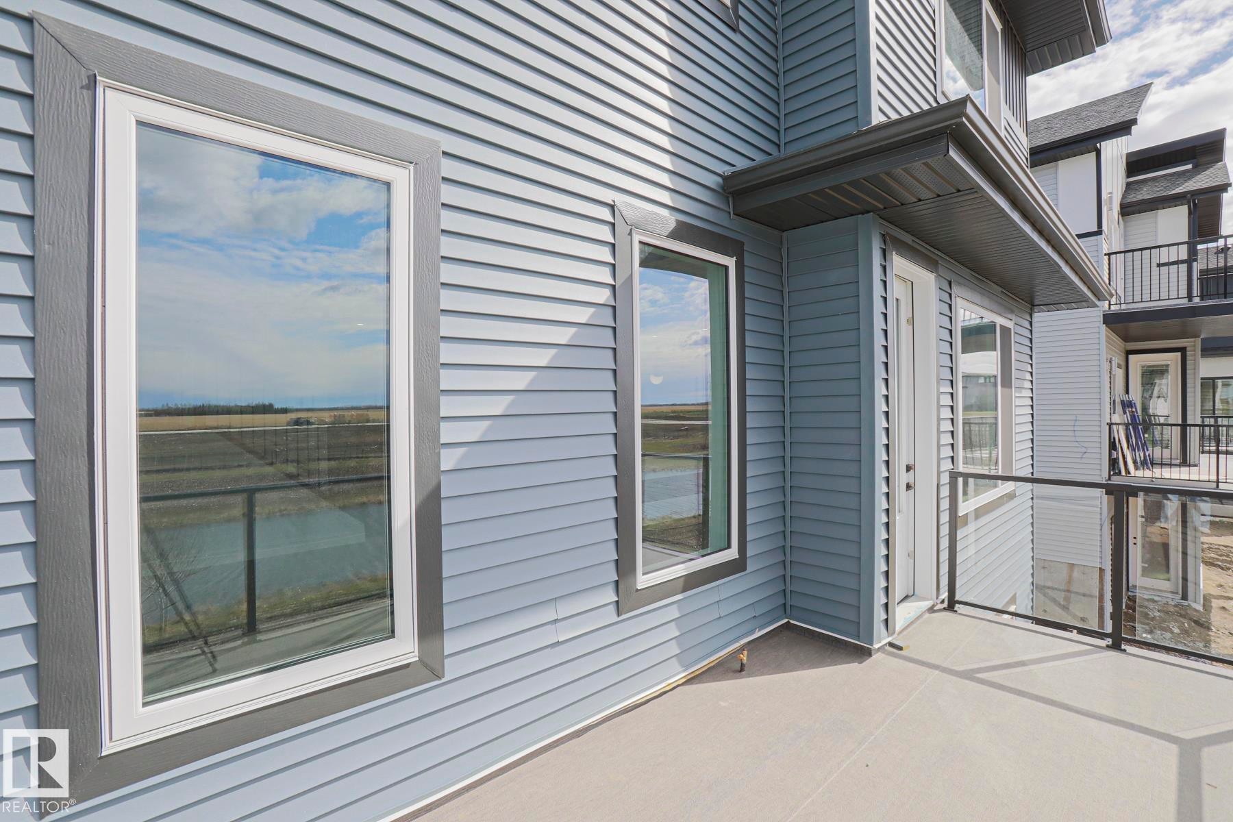 The property features light blue siding, a balcony with a glass railing, and a window reflecting a field and sky - Rural Leduc County, AB - Outdoor With Exterior
