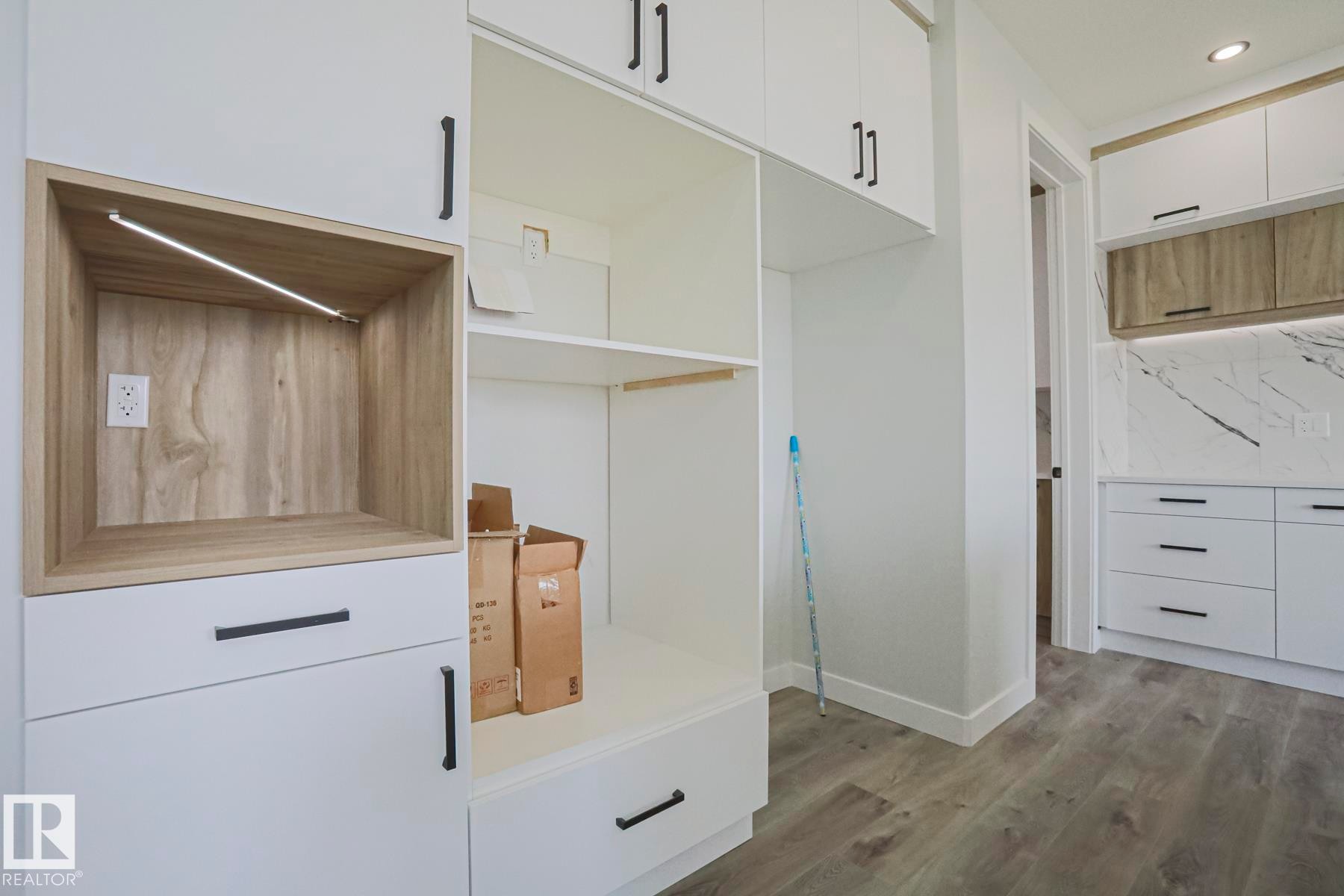 This space features contemporary white cabinetry with black hardware, a built-in wooden display niche with integrated lighting, and light wood-style flooring - Rural Leduc County, AB - Indoor