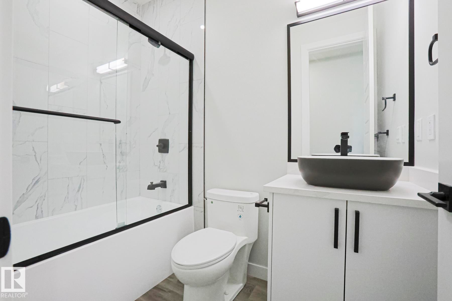 Bathroom featuring a white bathtub with a clear glass sliding shower door and black trim, a toilet, and a vanity with a dark gray vessel sink and black faucet - Rural Leduc County, AB - Indoor Photo Showing Bathroom
