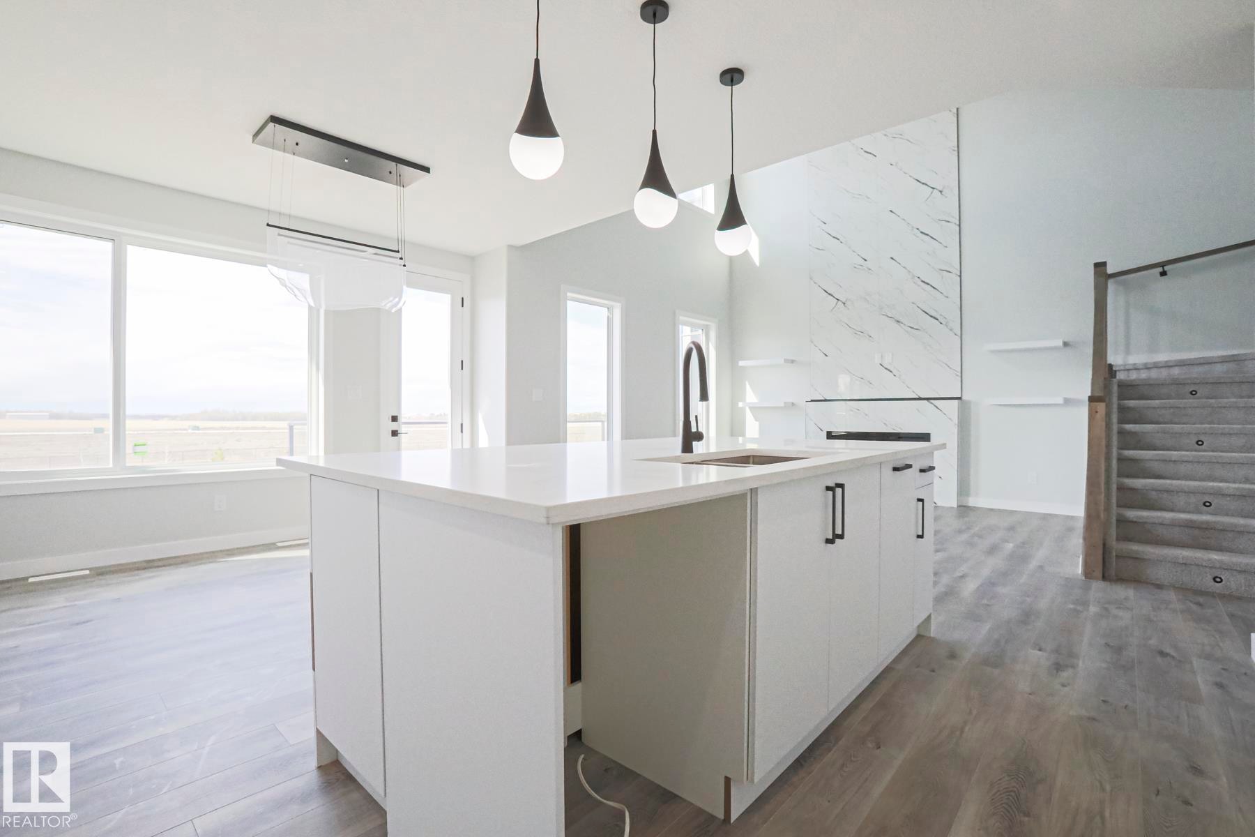 The open-concept living space features a kitchen island with a sink and modern pendant lighting, large windows, and a stone fireplace with shelving - Rural Leduc County, AB - Indoor Photo Showing Kitchen