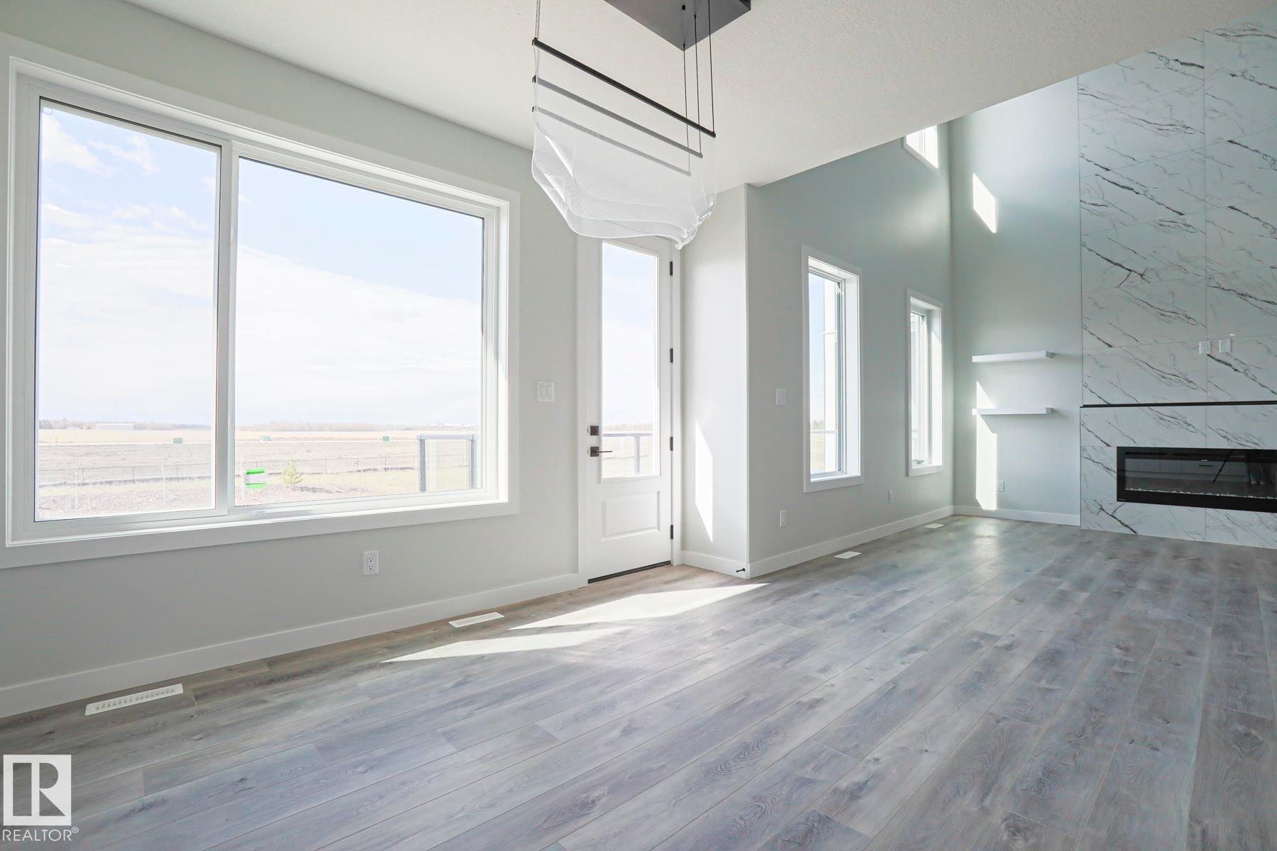 Living area featuring light grey flooring, large windows, a white door, and a fireplace with a stone surround - Rural Leduc County, AB - Indoor With Fireplace