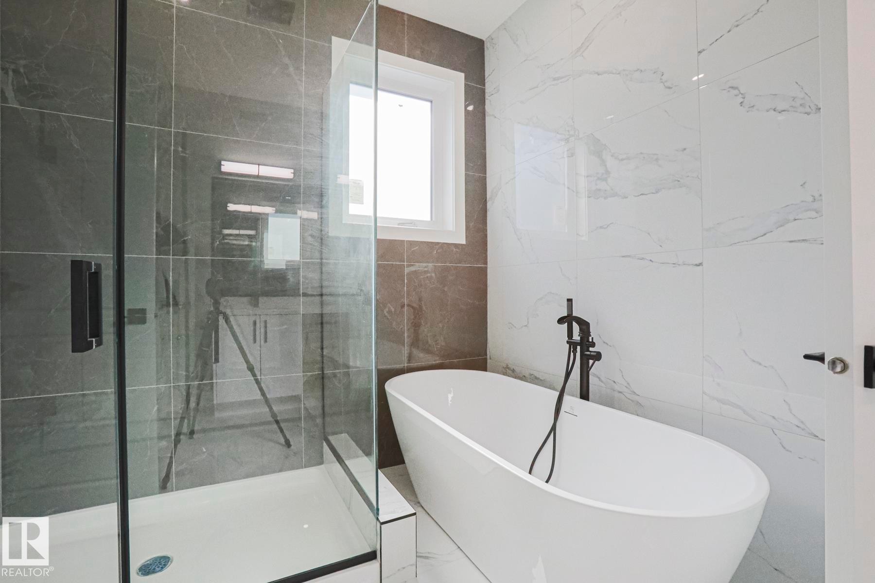The bathroom features a freestanding soaking tub with a matte black floor-mounted faucet, a glass-enclosed shower with a dark tiled wall, and white wall tiles with natural veining - Rural Leduc County, AB - Indoor Photo Showing Bathroom