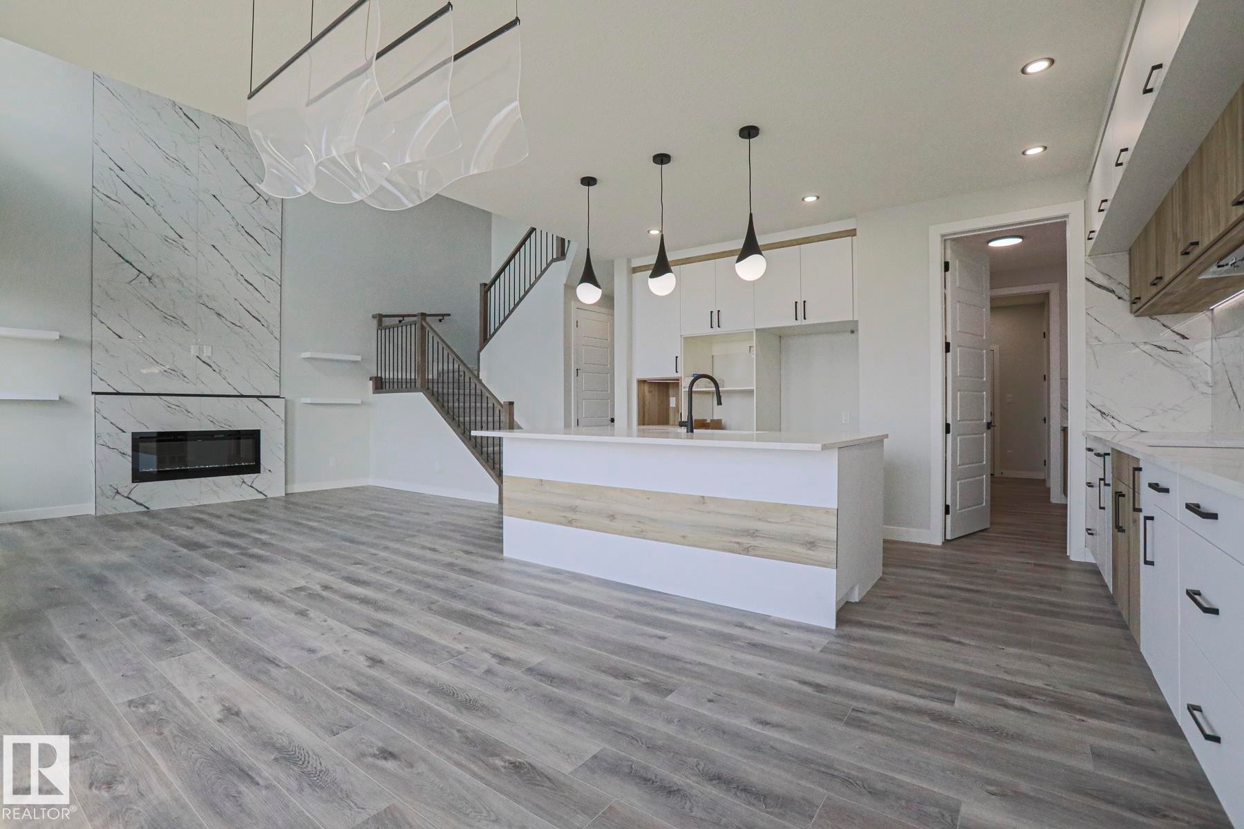 This expansive open-concept living area features light-toned flooring, a grand fireplace with a natural stone surround, and a kitchen island with pendant lighting - Rural Leduc County, AB - Indoor With Fireplace