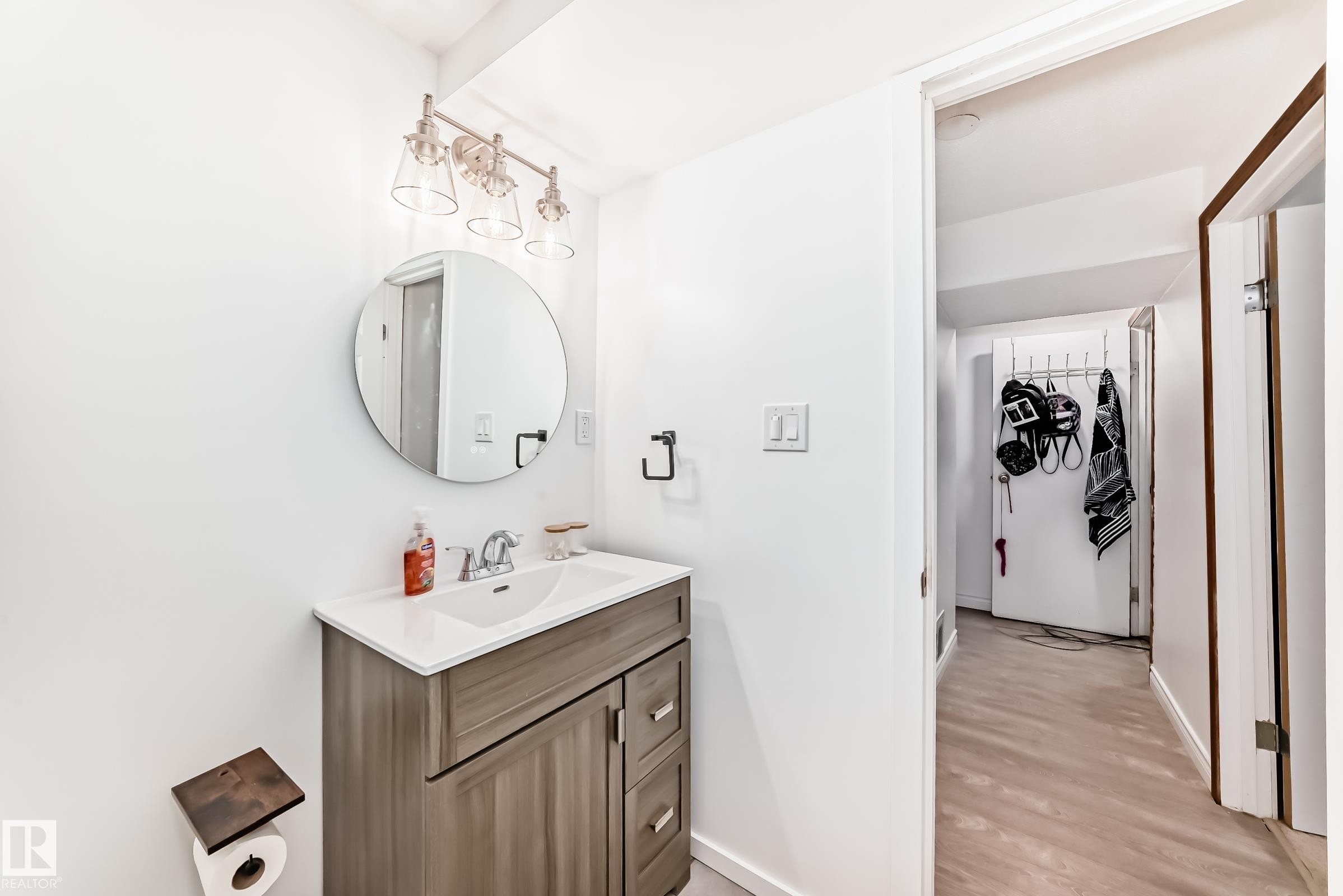The bathroom features a vanity with a white countertop and a circular mirror, illuminated by a modern light fixture with three clear glass shades - 11323 162 Avenue, Edmonton, AB - Indoor Photo Showing Bathroom