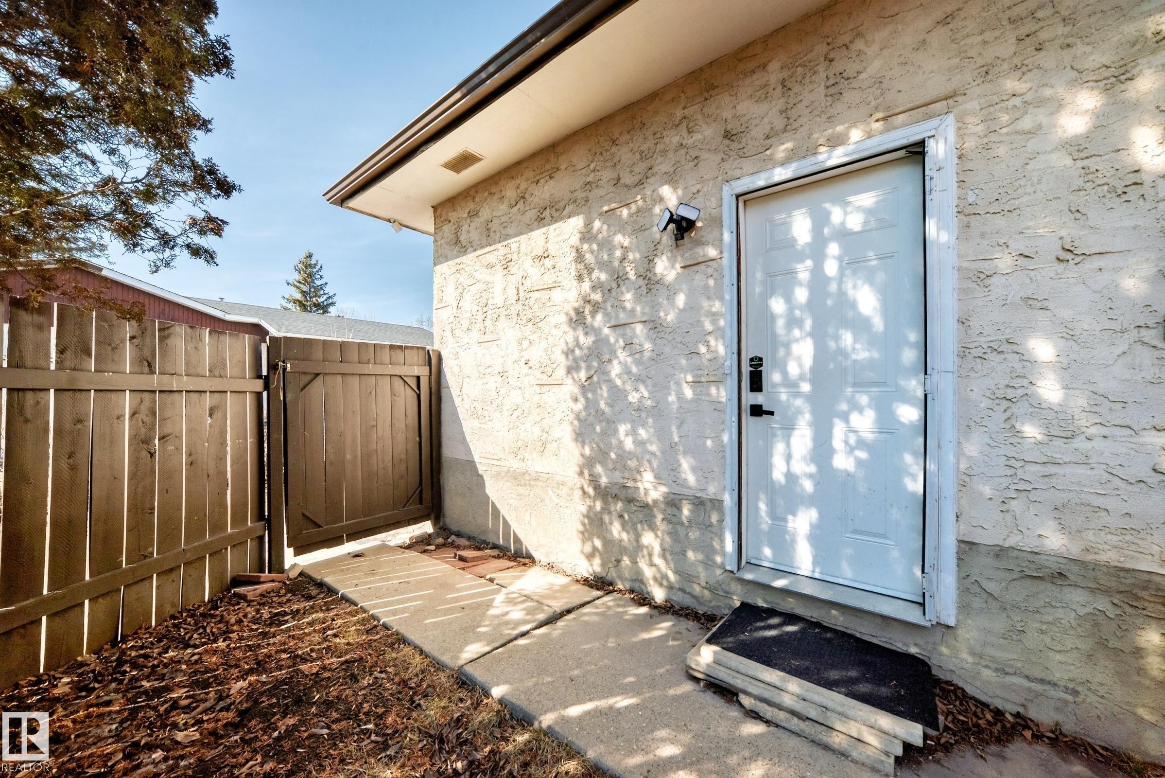 The exterior features a white door with dark hardware, a textured wall, and a concrete pathway leading to a wooden fence with a gate - 11323 162 Avenue, Edmonton, AB - Outdoor With Exterior