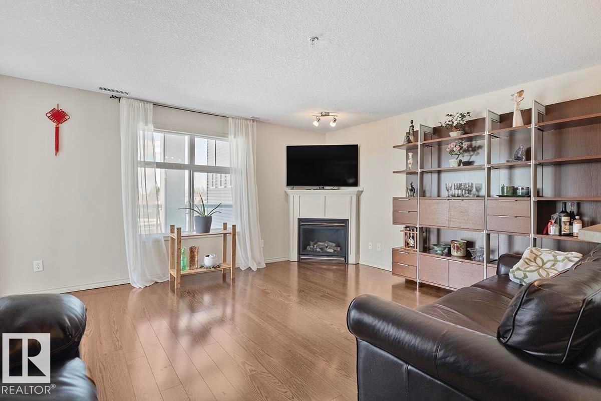 Living area featuring hardwood flooring, a gas fireplace with a mantel, and a large window with white curtains - 225 16035 132 Street, Edmonton, AB - Indoor Photo Showing Living Room With Fireplace