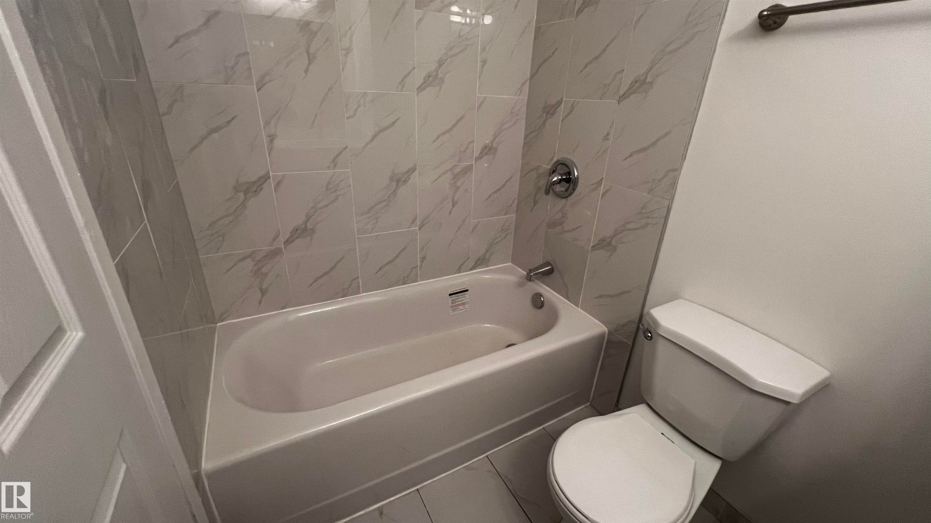 The bathroom features a bathtub with a shower, a toilet, and light-colored wall tiles with a subtle marble pattern - 3323 107 Avenue Nw, Edmonton, AB - Indoor Photo Showing Bathroom