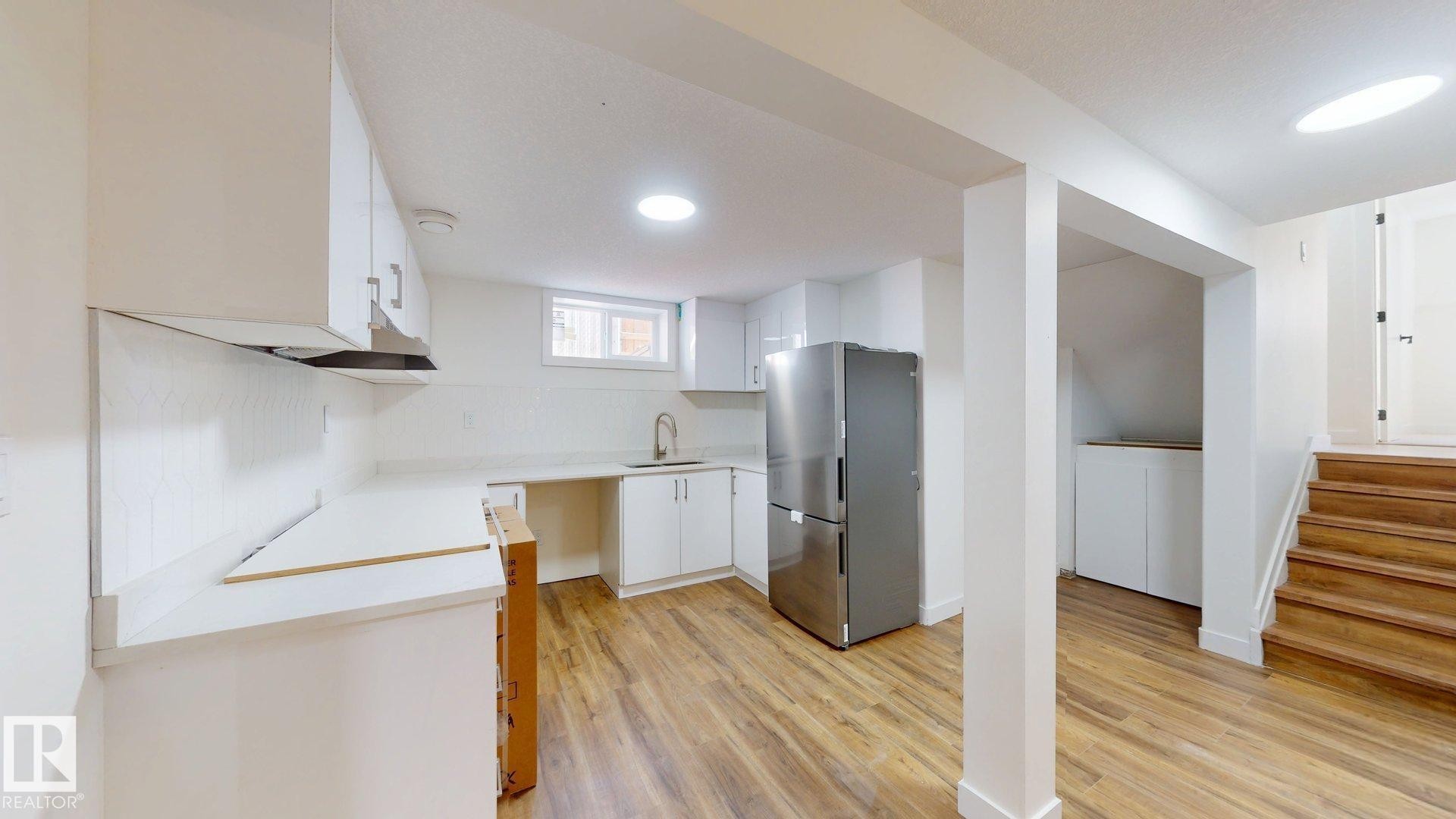 This property features a kitchen area with white cabinetry, a stainless steel refrigerator, and wood-look flooring - 8515 86 Avenue, Edmonton, AB - Indoor