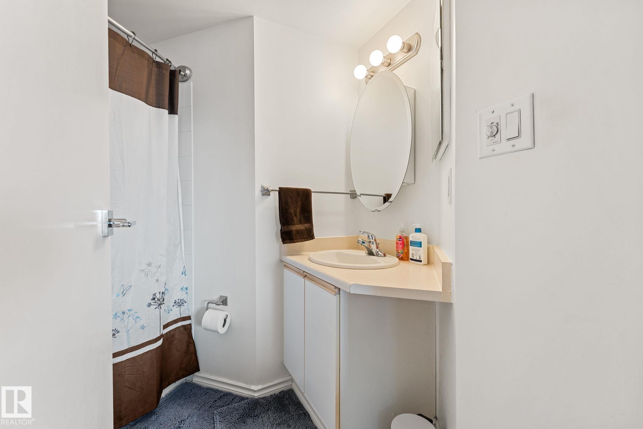 The bathroom features a vanity with a countertop and sink, an oval mirror, and a shower area - 208 6710 158 Avenue, Edmonton, AB