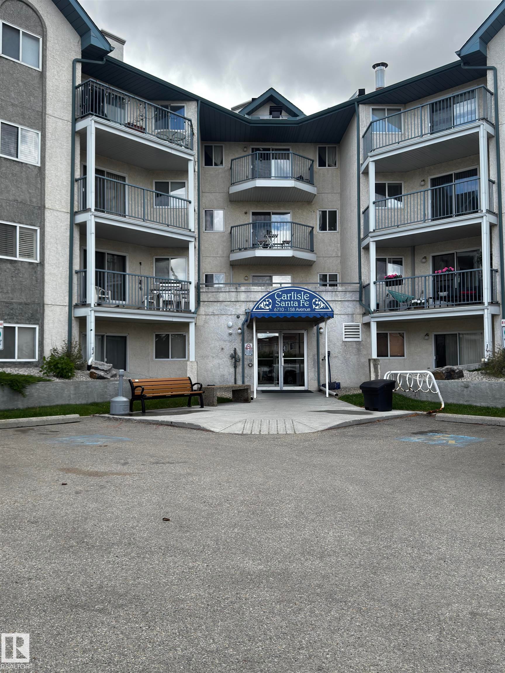 Multi-story residential building featuring multiple balconies, a central entrance with a blue awning, and an asphalt parking area - 208 6710 158 Avenue, Edmonton, AB
