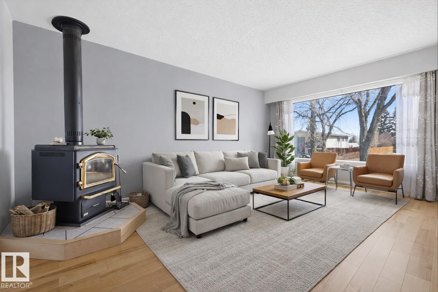 Spacious living area featuring light wood flooring, a large window providing natural light, and a black wood-burning stove with a gold-toned door - 10612 79 Street, Edmonton, AB