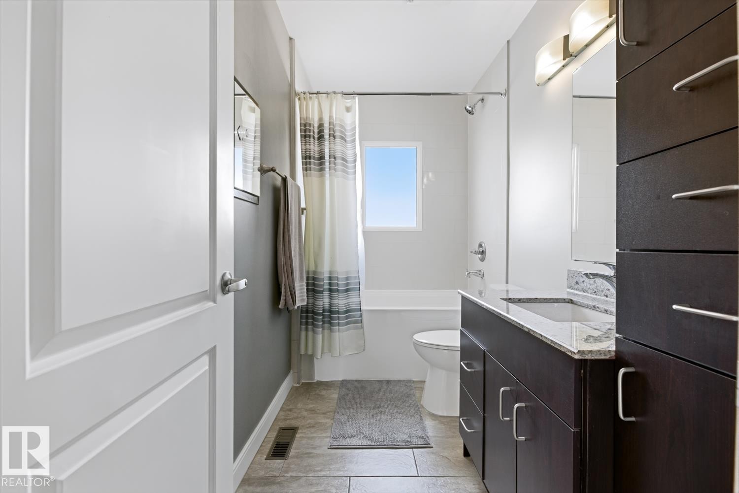 Bathroom featuring a dark wood vanity with a granite countertop, a built-in bathtub with a shower, and light-colored tile flooring - 10612 79 Street, Edmonton, AB