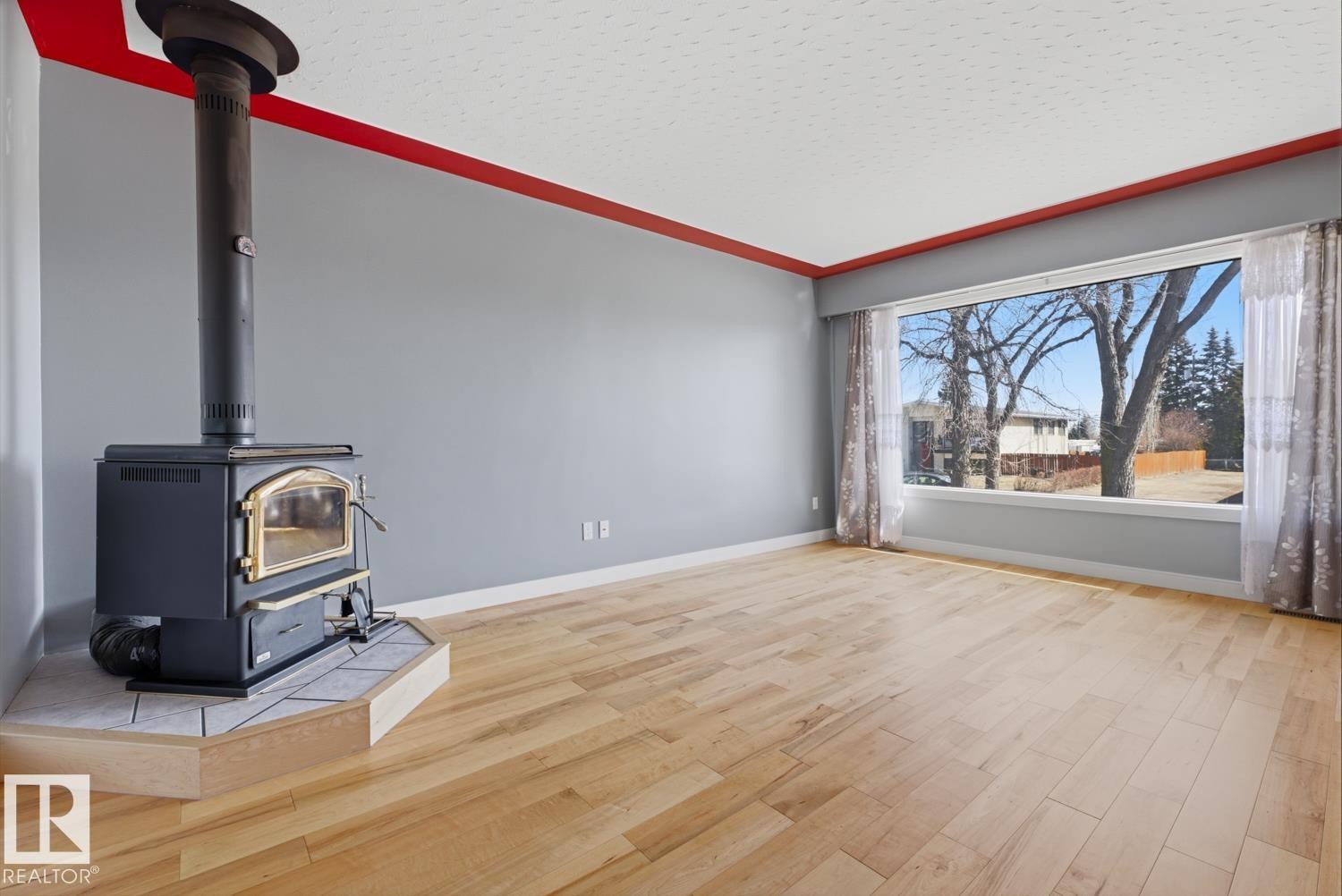 Living area featuring light wood flooring, a corner wood-burning stove with a tiled base, and a large window providing views of trees and other properties - 10612 79 Street, Edmonton, AB