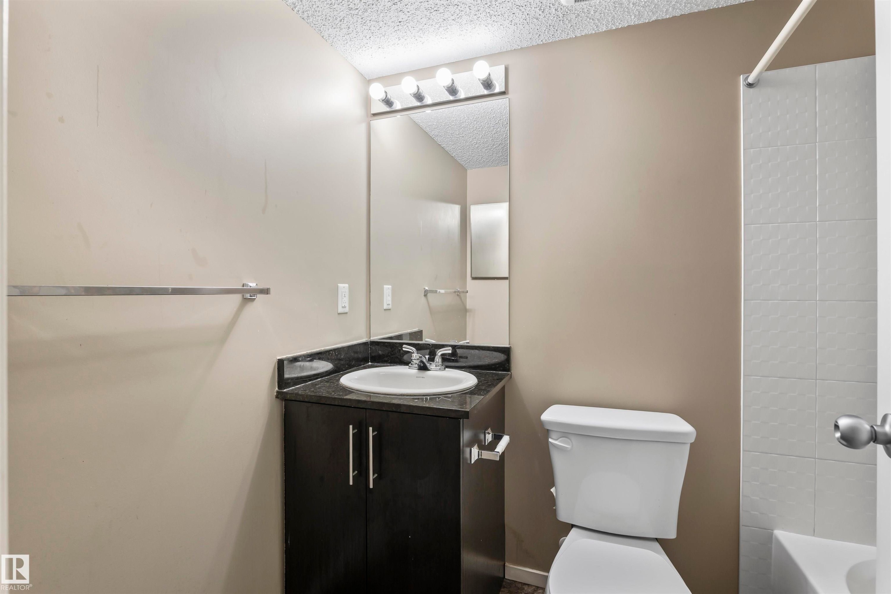 The bathroom features a vanity with a dark countertop and an integrated sink, a large mirror with overhead lighting, and a white toilet - 214 5816 Mullen Place, Edmonton, AB - Indoor Photo Showing Bathroom