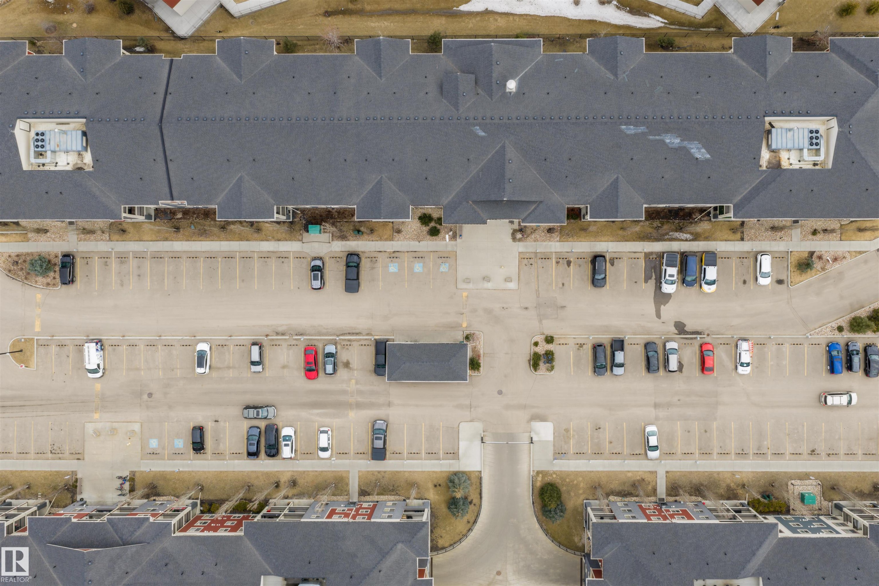 This aerial view showcases the property’s expansive paved parking areas and the well-maintained dark grey shingled roofs of the buildings - 214 5816 Mullen Place, Edmonton, AB - Photo Showing Garage