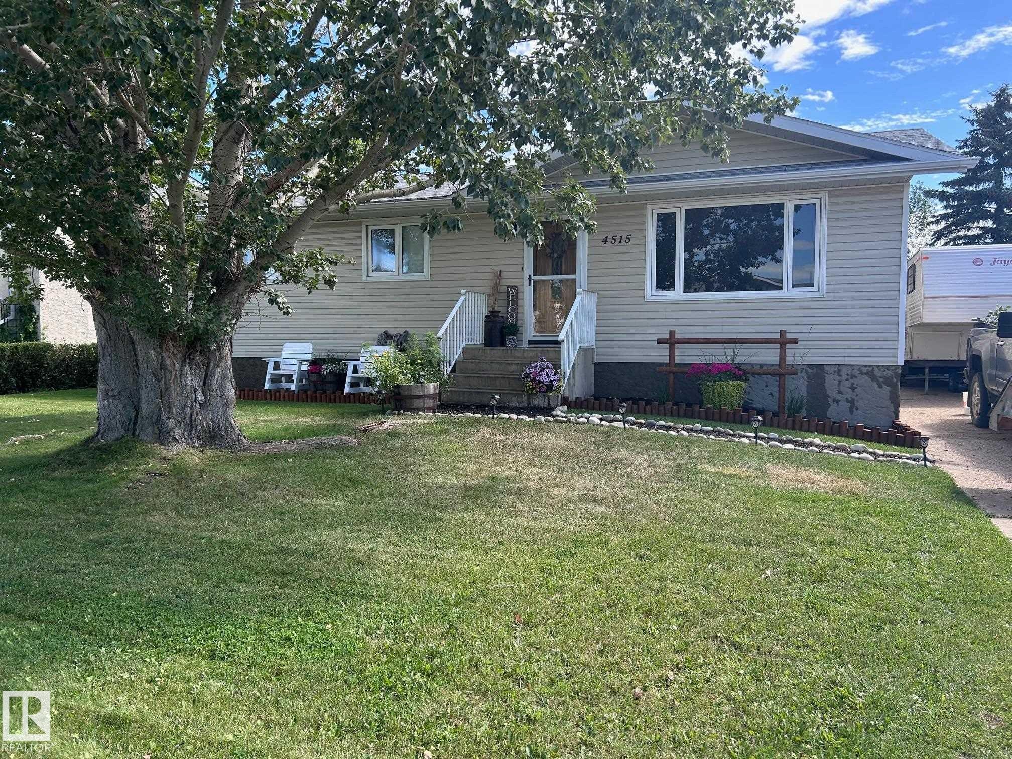 Exterior view of the property featuring light-colored siding, a front yard with a mature tree, and a paved driveway - 4515 54 Ave, Tofield, AB - Outdoor