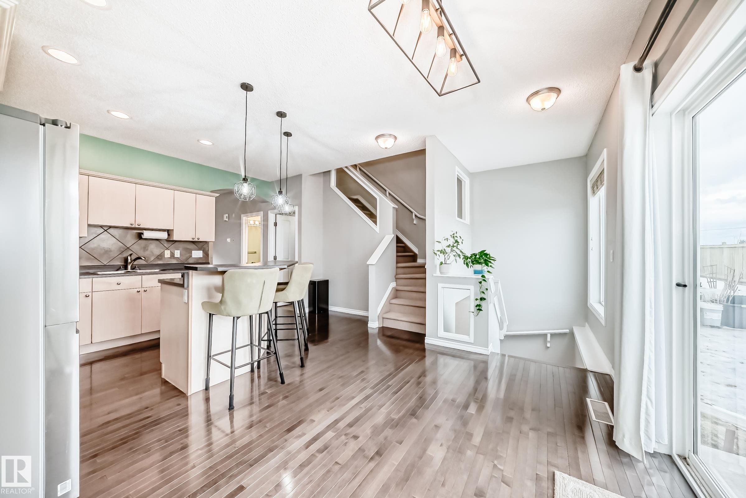 The open concept living area features hardwood floors, a kitchen island with pendant lighting, a stainless steel refrigerator, and a sliding glass door leading to an outdoor space - 105 13825 155 Avenue, Edmonton, AB - Indoor Photo Showing Kitchen With Upgraded Kitchen