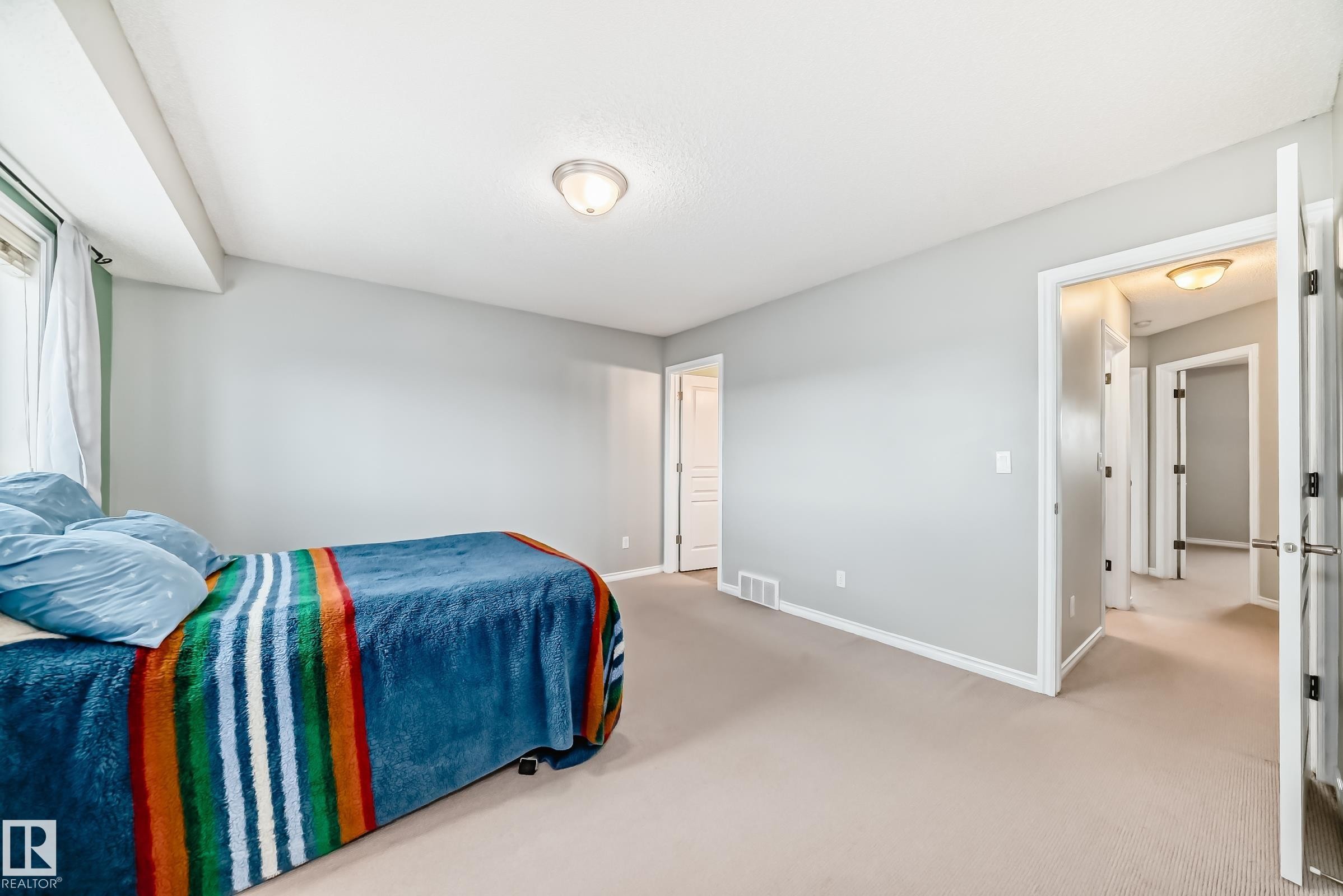 Spacious room featuring light-colored carpet, light grey walls, and a window with white curtains - 105 13825 155 Avenue, Edmonton, AB - Indoor Photo Showing Bedroom