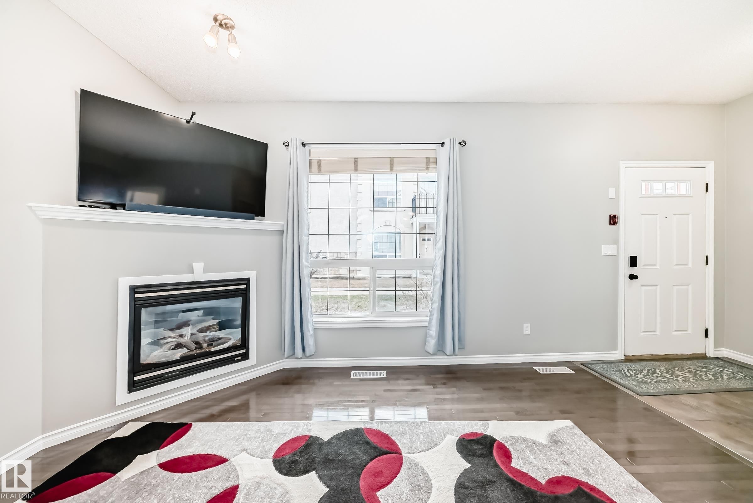 The property features hardwood floors, a prominent white door with glass panels, and a window with light blue curtains - 105 13825 155 Avenue, Edmonton, AB - Indoor Photo Showing Living Room With Fireplace