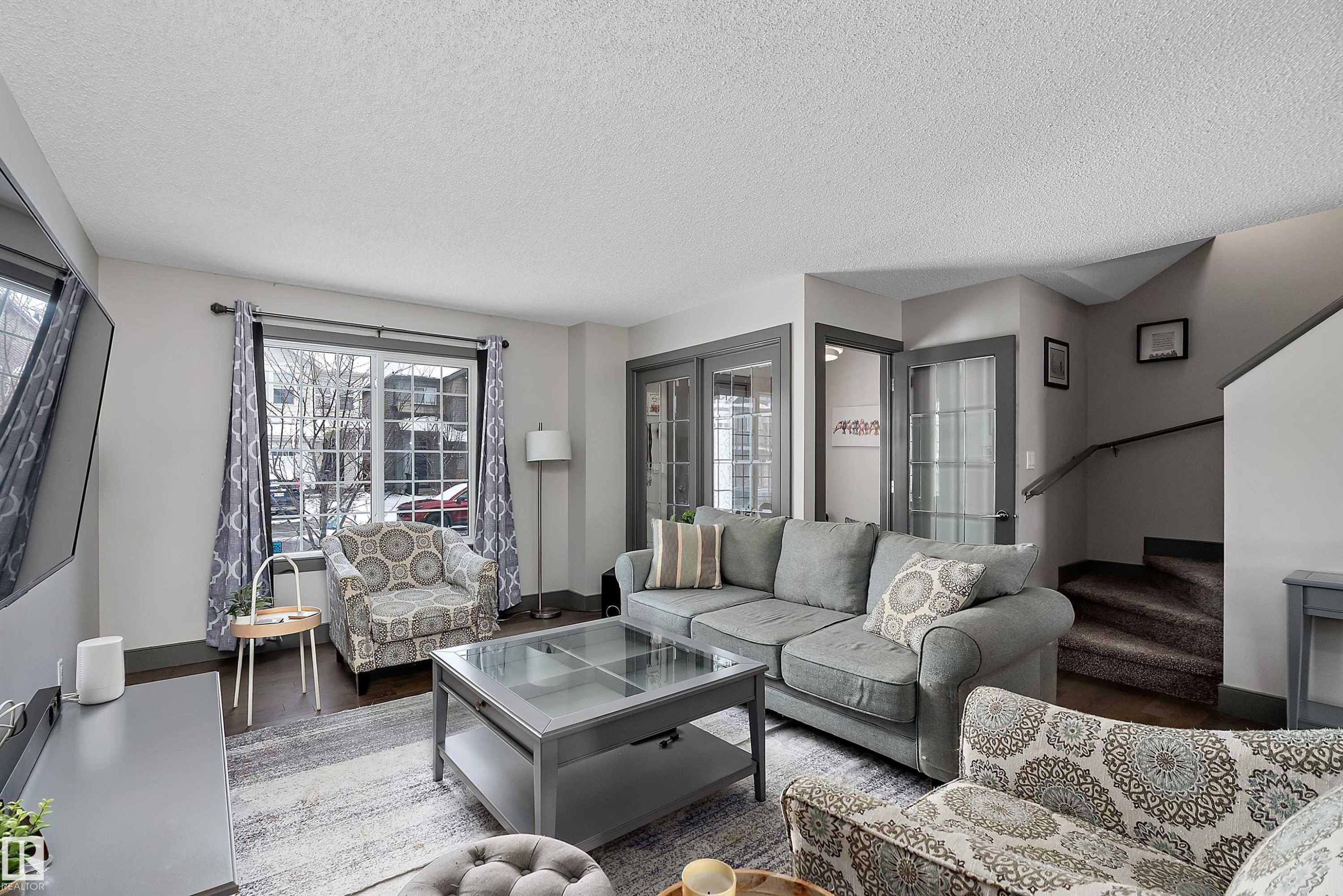 Well-appointed living area featuring hardwood flooring, a large window with white grilles, and a staircase with a dark handrail - 7776 Eifert Crescent Nw, Edmonton, AB - Indoor Photo Showing Living Room